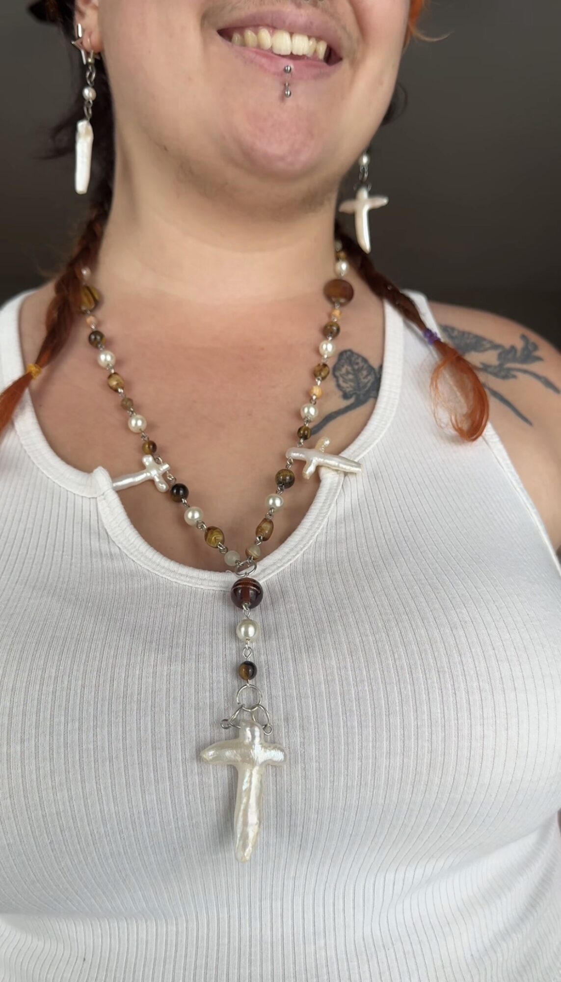 Person wearing a white tank top with a cross necklace, earrings, and a tattoo on shoulder, smiling.