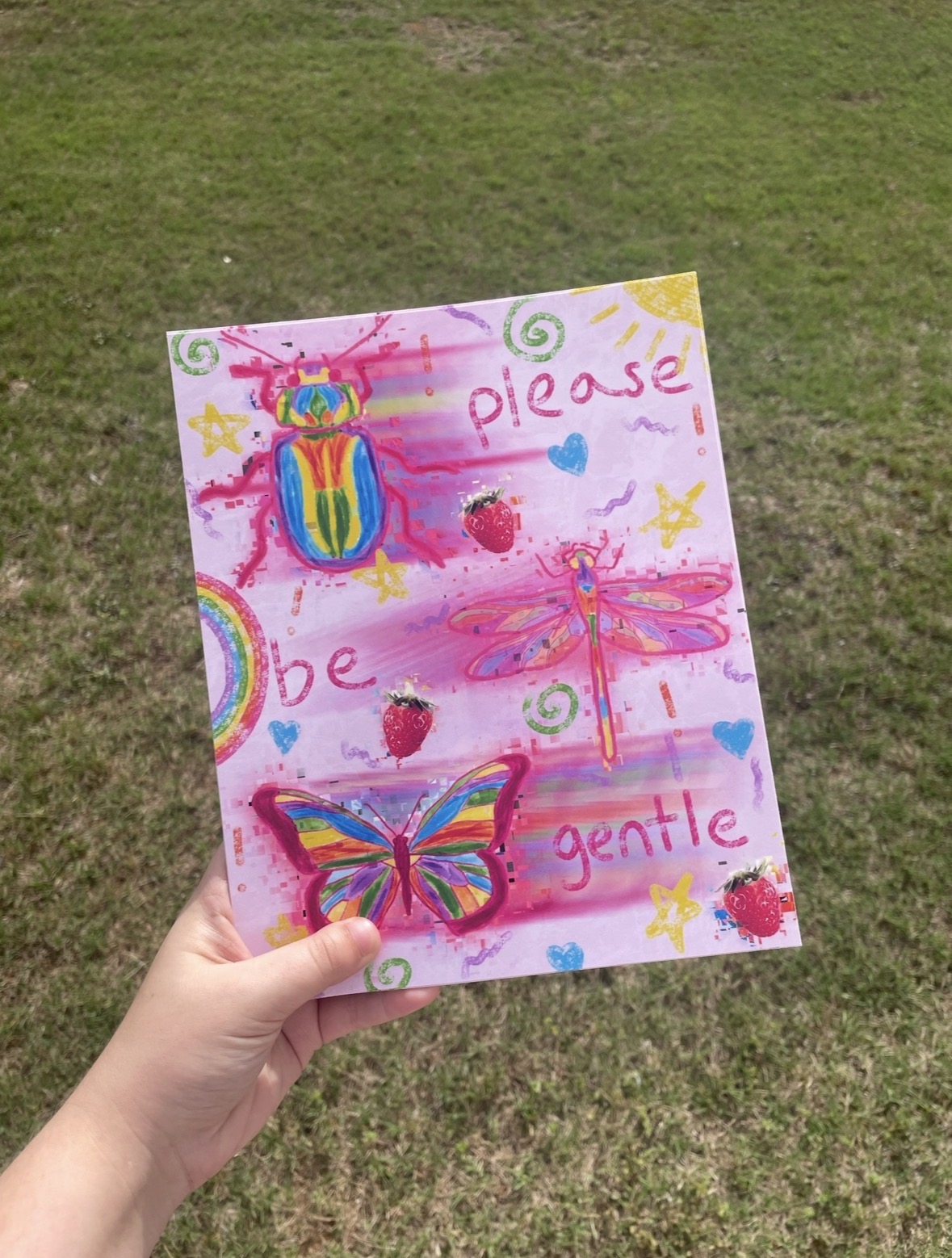 Hand holding a colorful, painted sign with a ladybug, butterfly, and dragonfly, on a grassy background, with words 'please be gentle'.