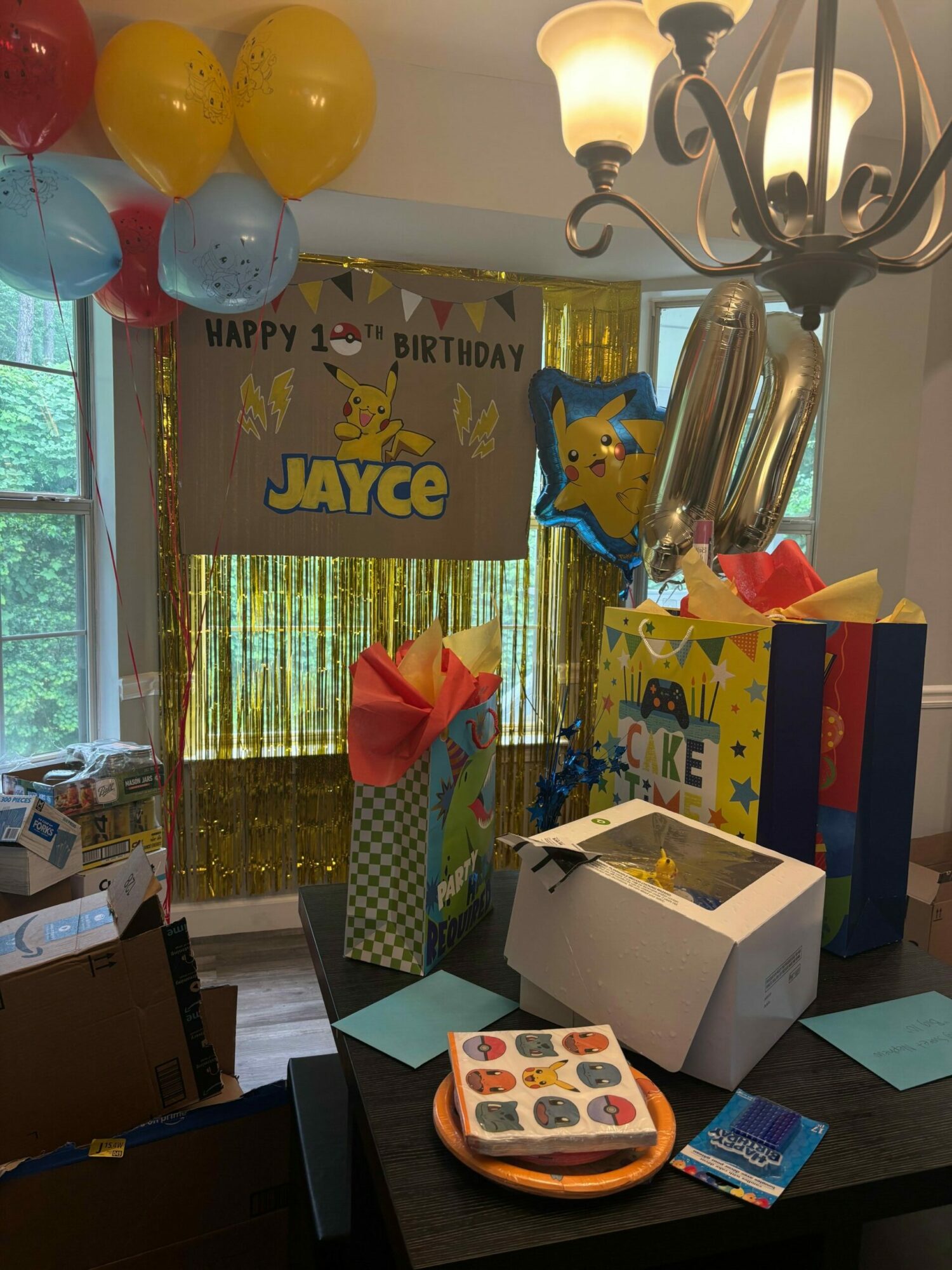 Party table with gifts, balloons, and decorations, in front of a window with curtains, celebrating a birthday.