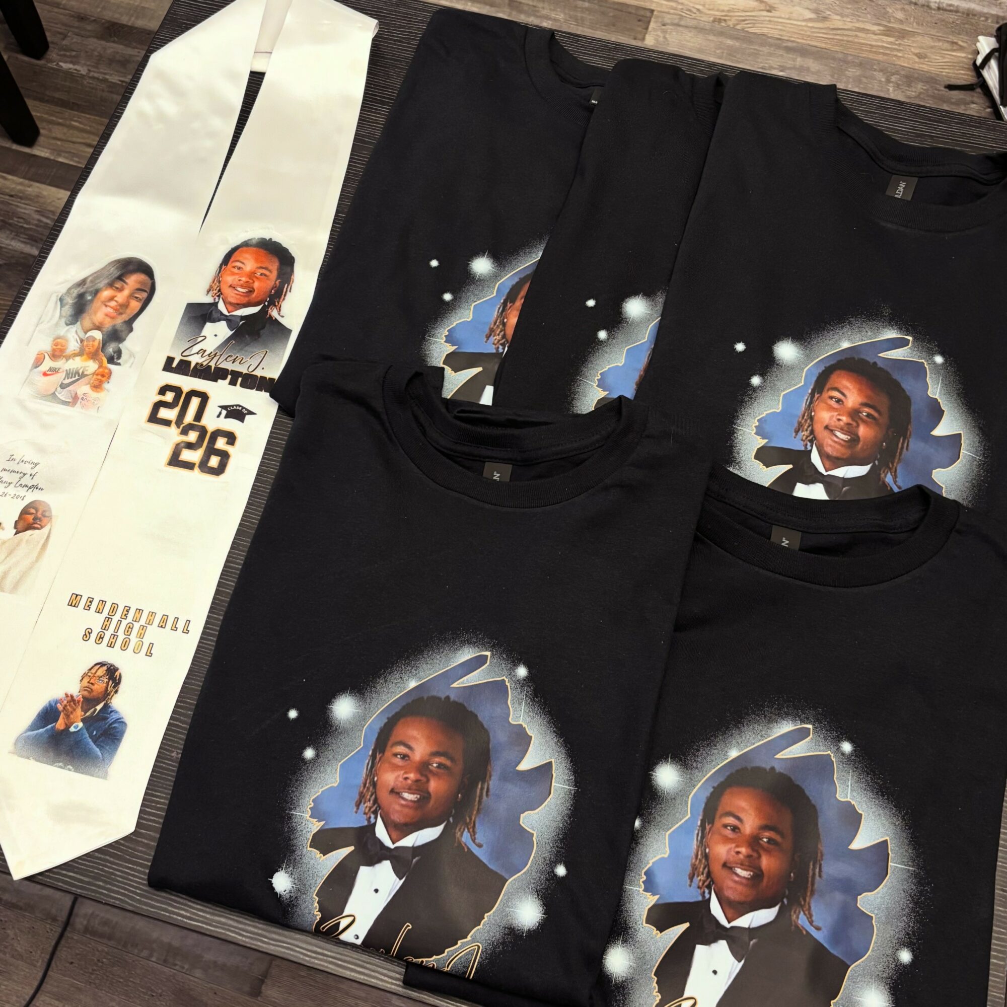 Four black T-shirts with a smiling girl in a tuxedo, surrounded by a starry background, laid on a wooden surface.