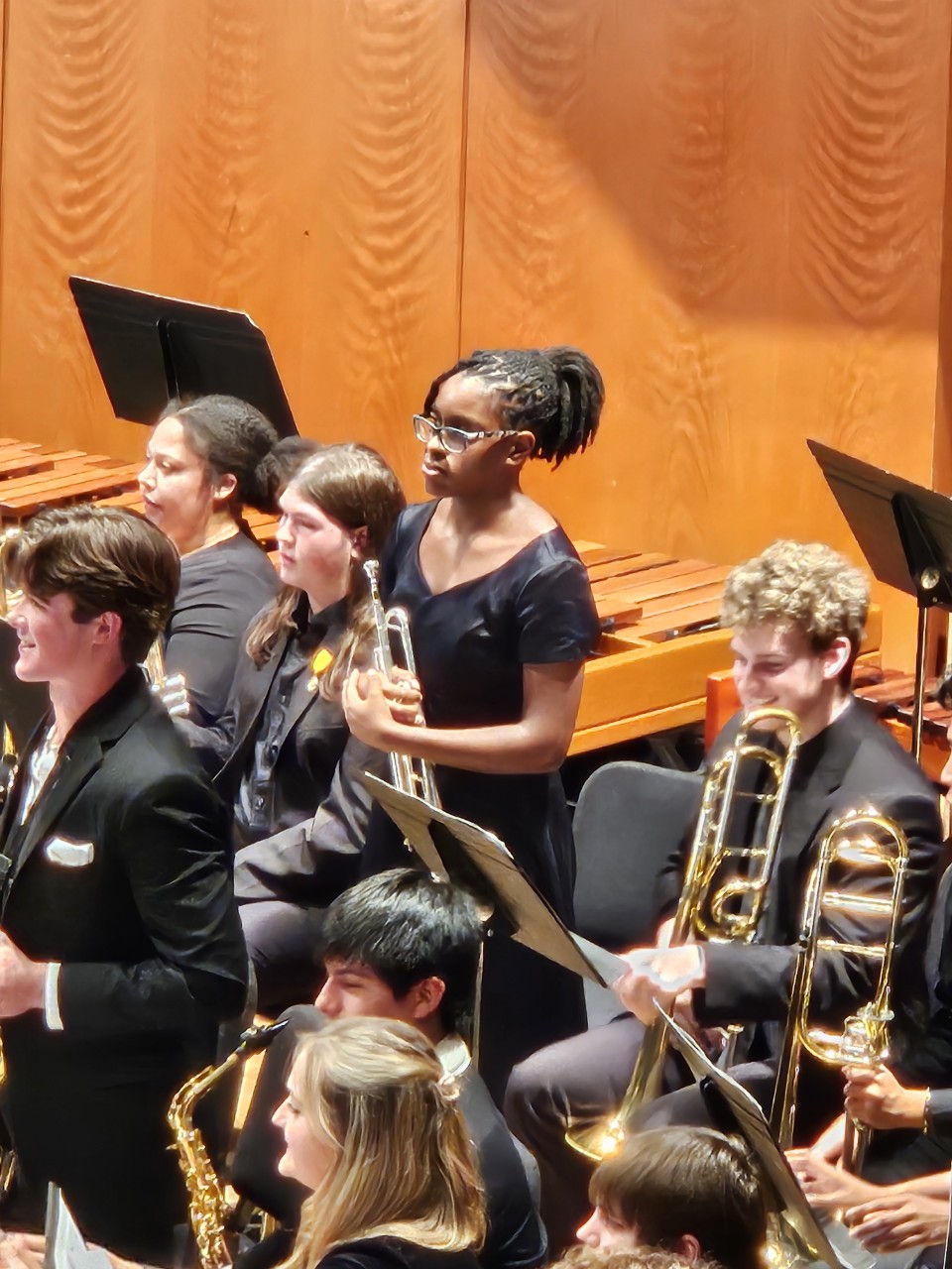 Group of young musicians in formal attire holding instruments on stage with orange curtains behind them.
