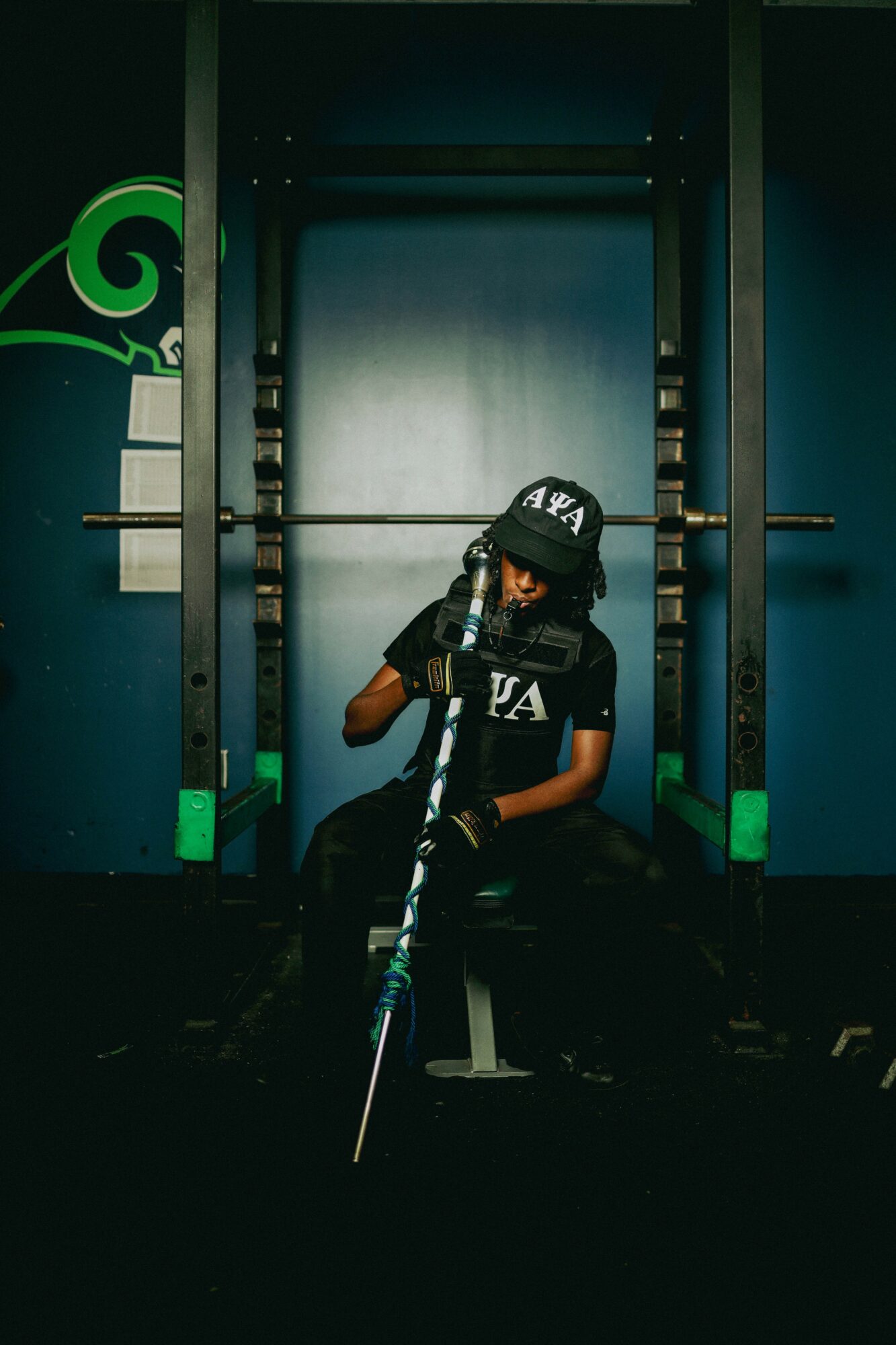 Person sitting in front of a large metal door, holding a hockey stick, wearing a cap and gloves, in a dark room.