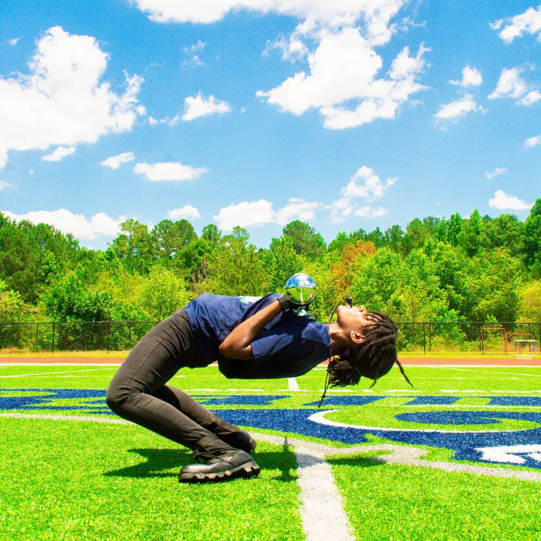 Person crouching on grass field holding a ball, with trees and blue sky in background.