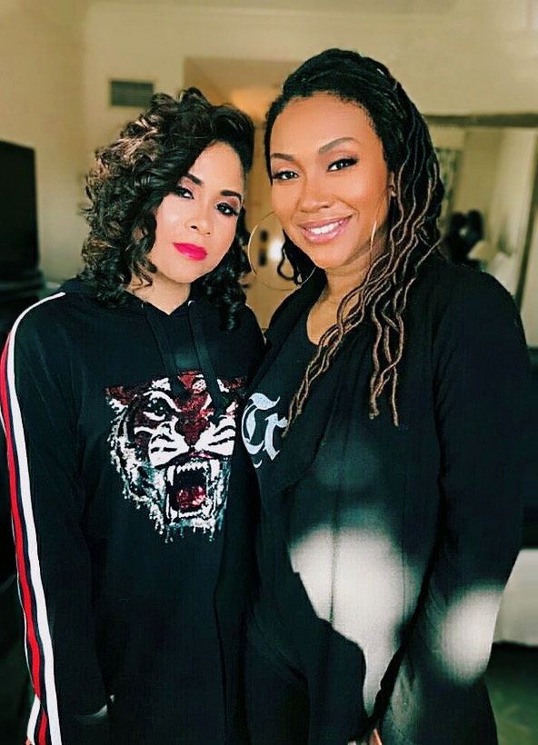 Two women smiling, one with curly hair and red lipstick, the other with dreadlocks, wearing dark clothing, indoors.