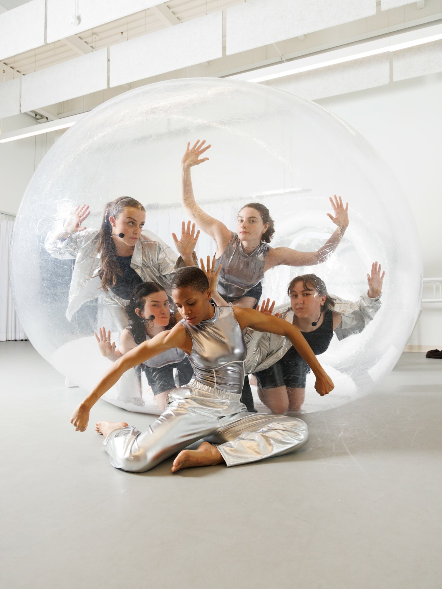 Five dancers inside a transparent bubble, posing with arms raised and extended, in a modern studio setting.