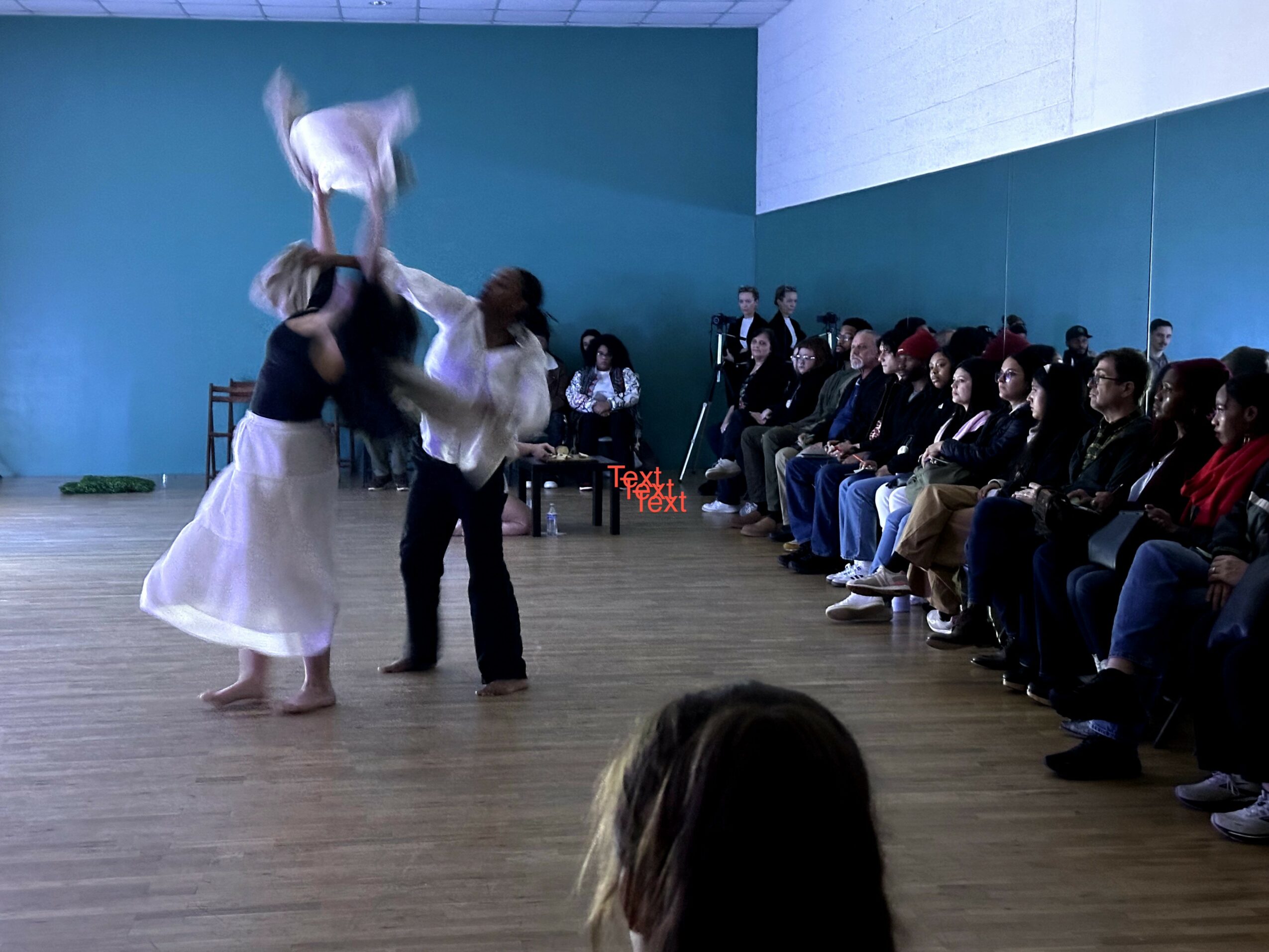 Two dancers perform in front of seated audience in a spacious room with blue walls.