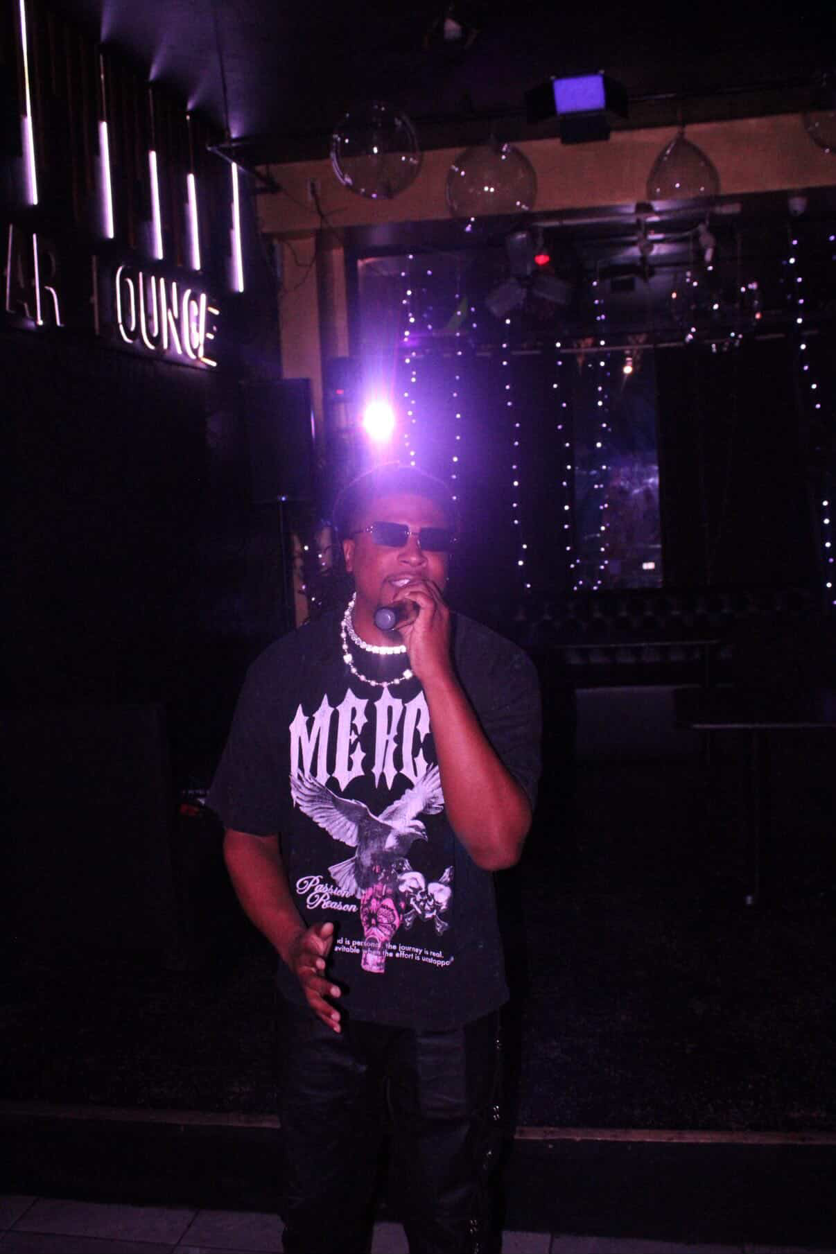 Person standing in a dimly lit venue with purple and blue lighting, wearing sunglasses, a graphic t-shirt, and jewelry.