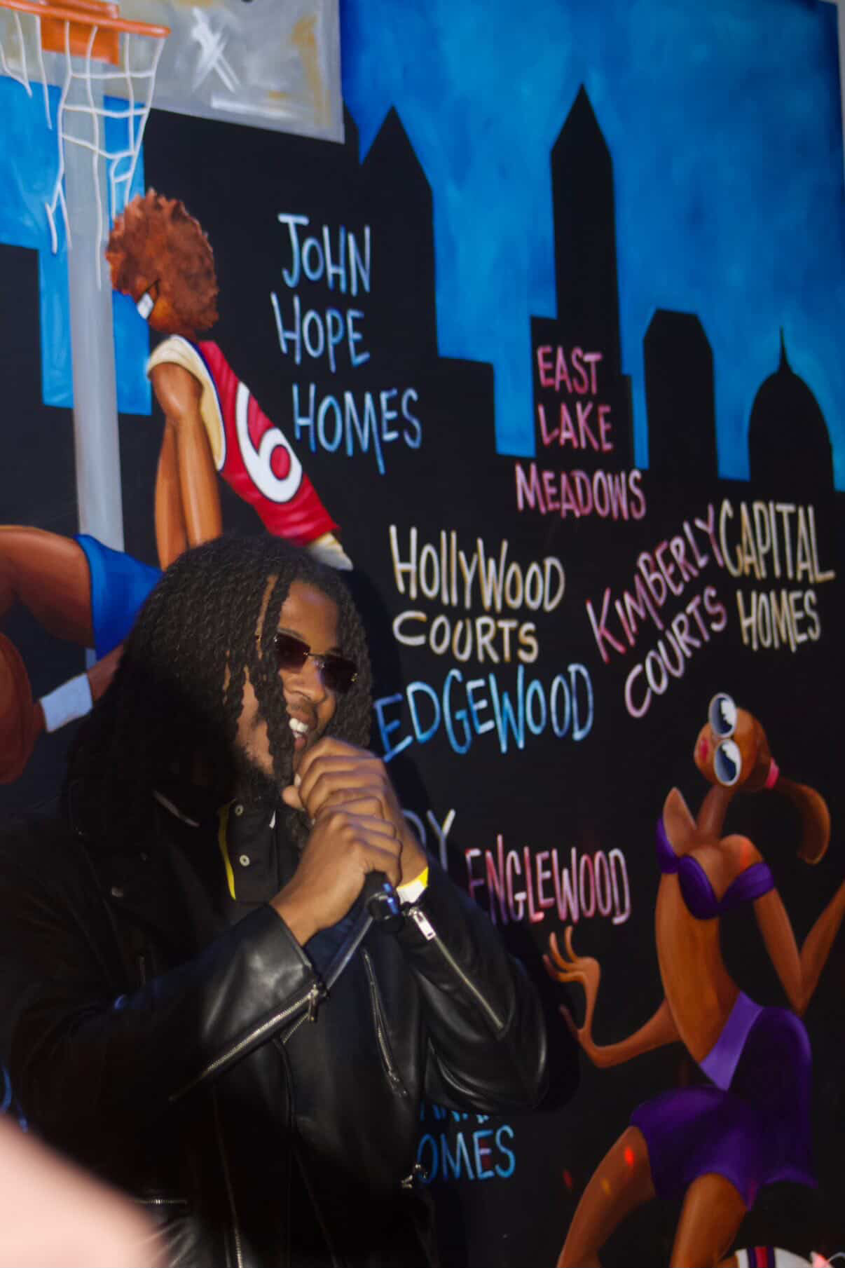 Person with sunglasses and leather jacket in front of a colorful cityscape mural with cartoon characters and city names.