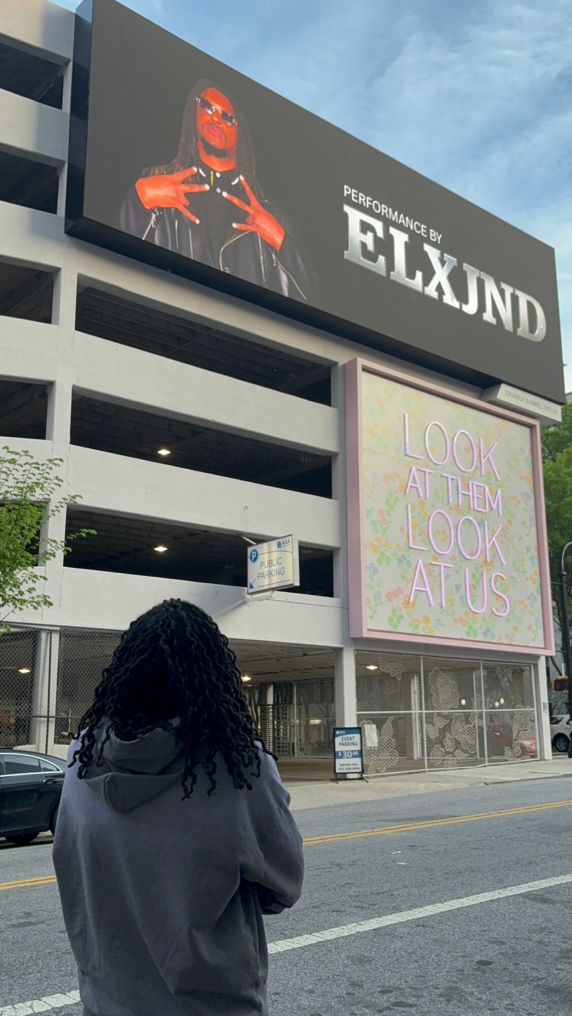 Person with curly hair and gray hoodie standing on street, looking at large digital billboards on building.