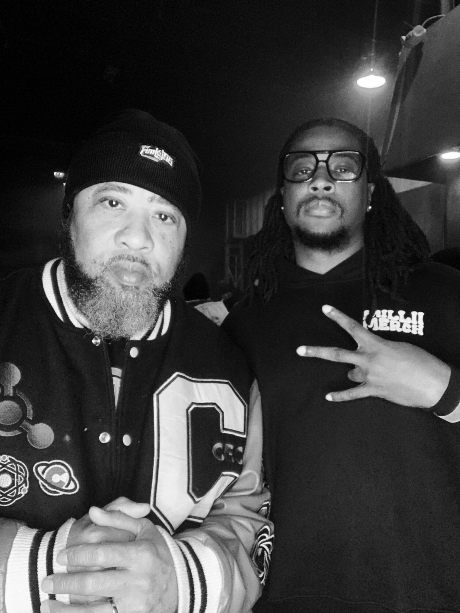 Two men pose indoors, one wearing a beanie and varsity jacket, the other with glasses and dreadlocks, making a peace sign.