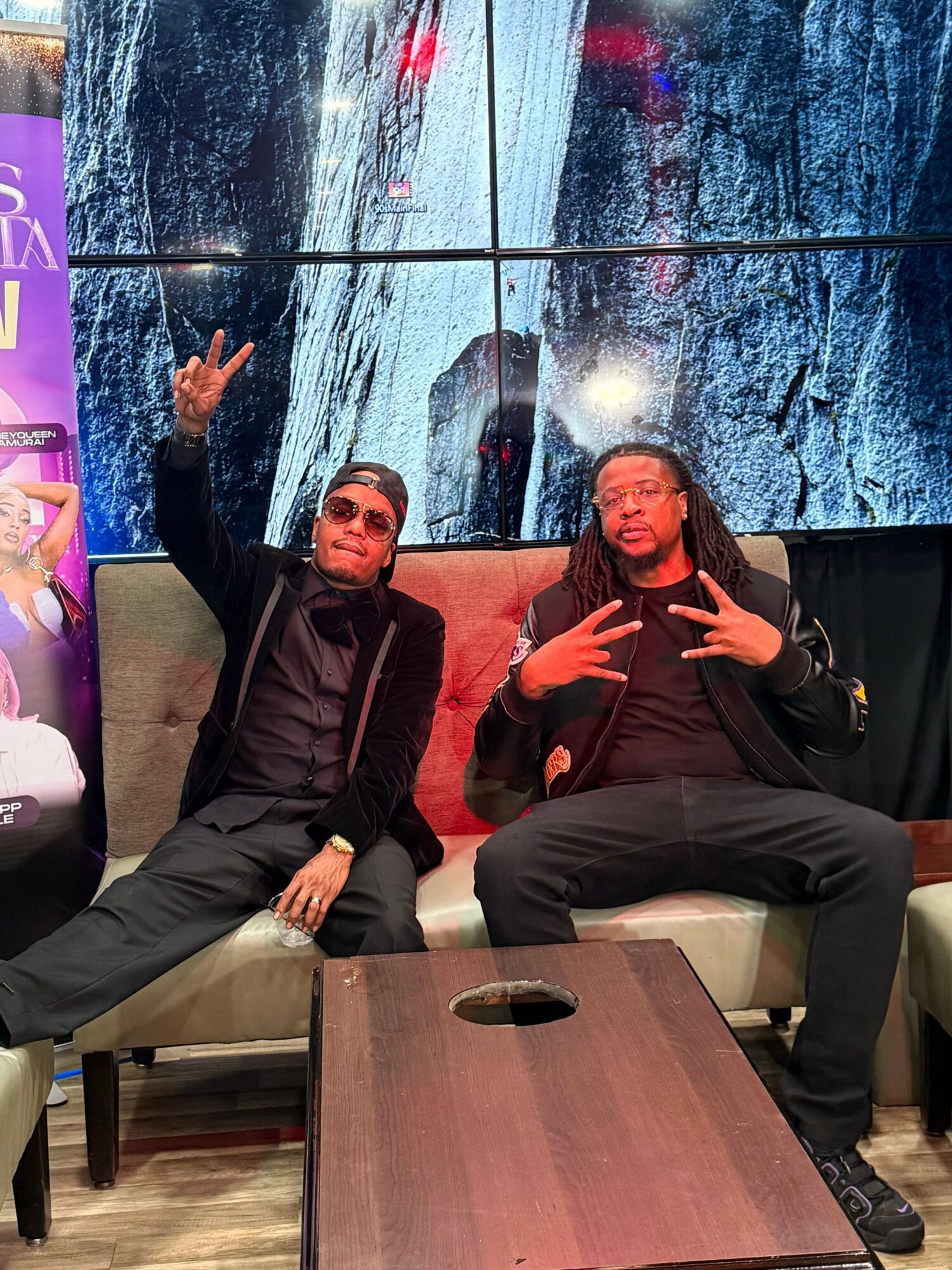 Two men sitting indoors near a large window with a snowy landscape outside, one making a peace sign and the other resting his hand on his knee.