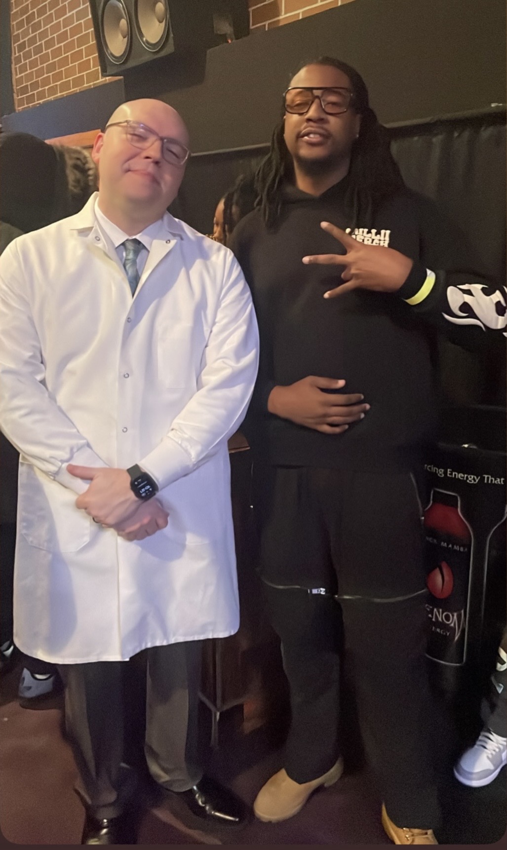 Two men standing indoors, one in a white lab coat and the other in a black hoodie, smiling and posing for the photo.