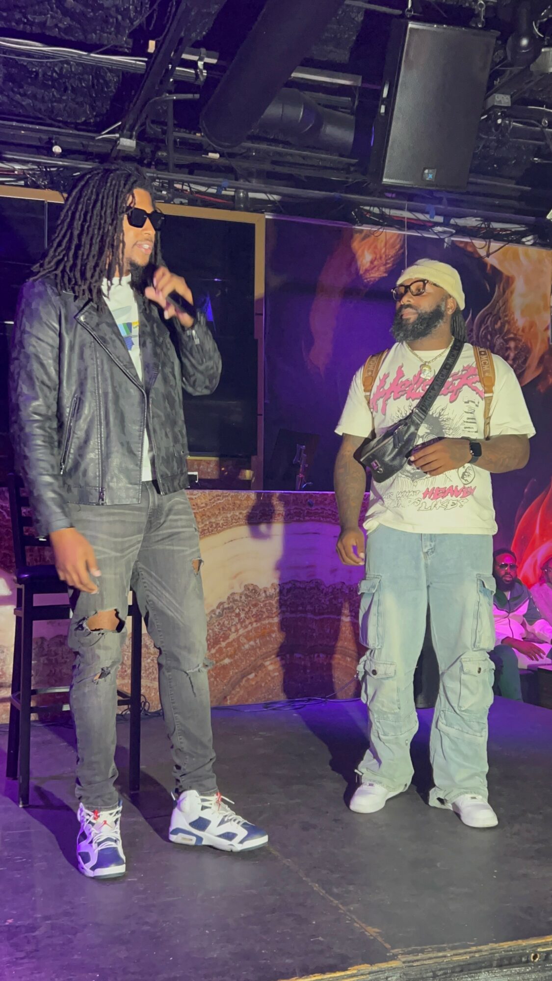 Two men stand on stage with a colorful background, one holding a microphone and the other with a bag, both wearing casual clothing.