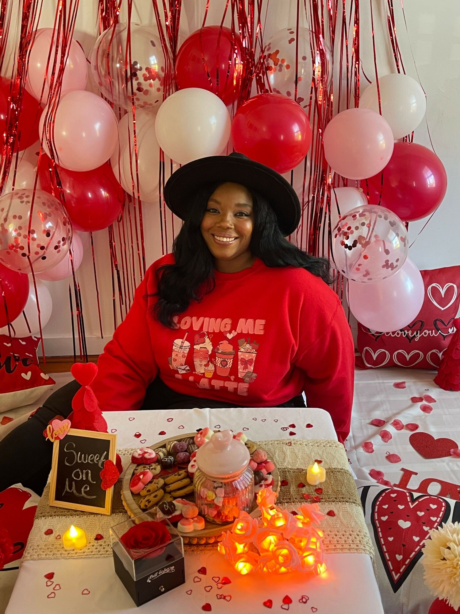 Person in a red hoodie and black hat sitting at a table with balloons and a cake, celebrating a special occasion.