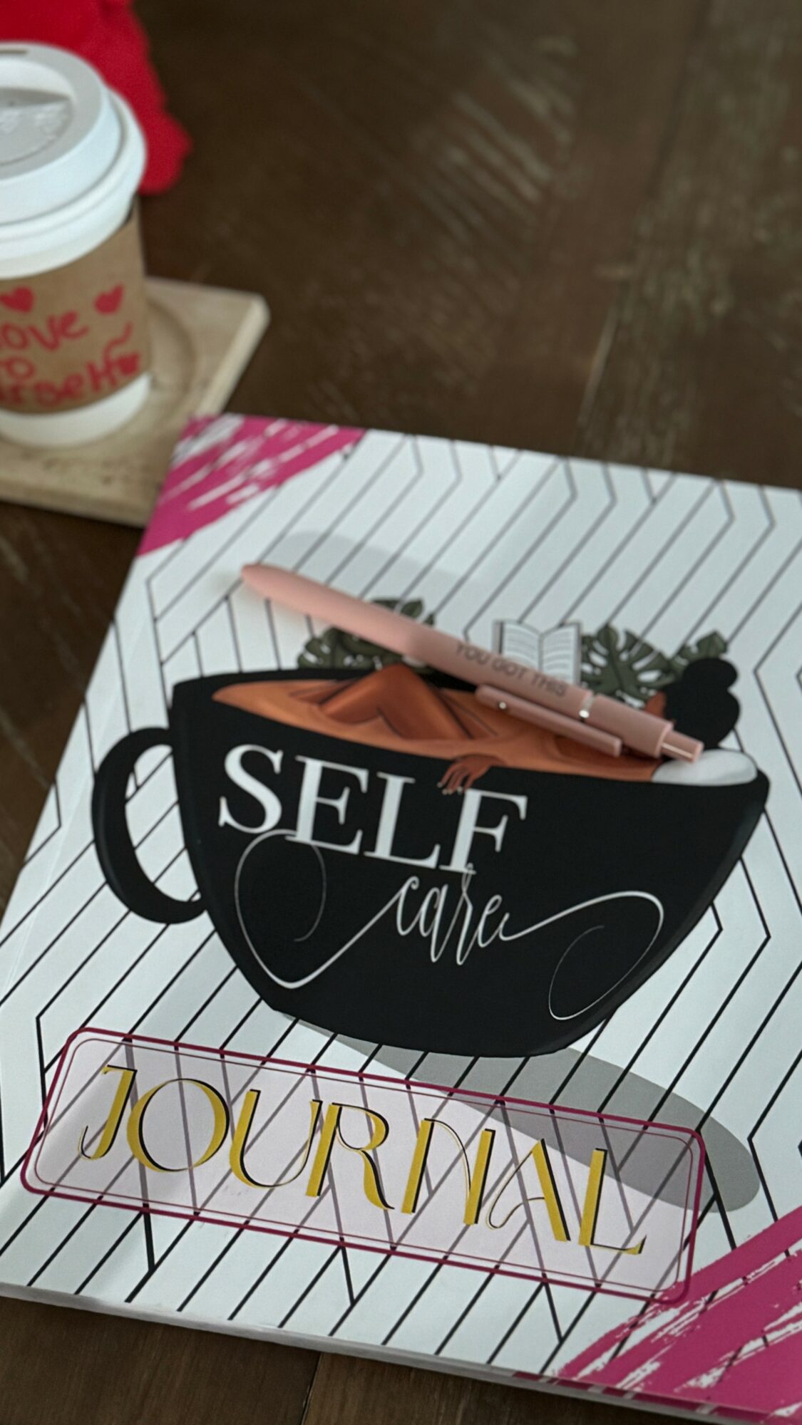 A notebook with a black coffee cup illustration and the words 'SELF CARE' on the cover, with a pen resting on it.