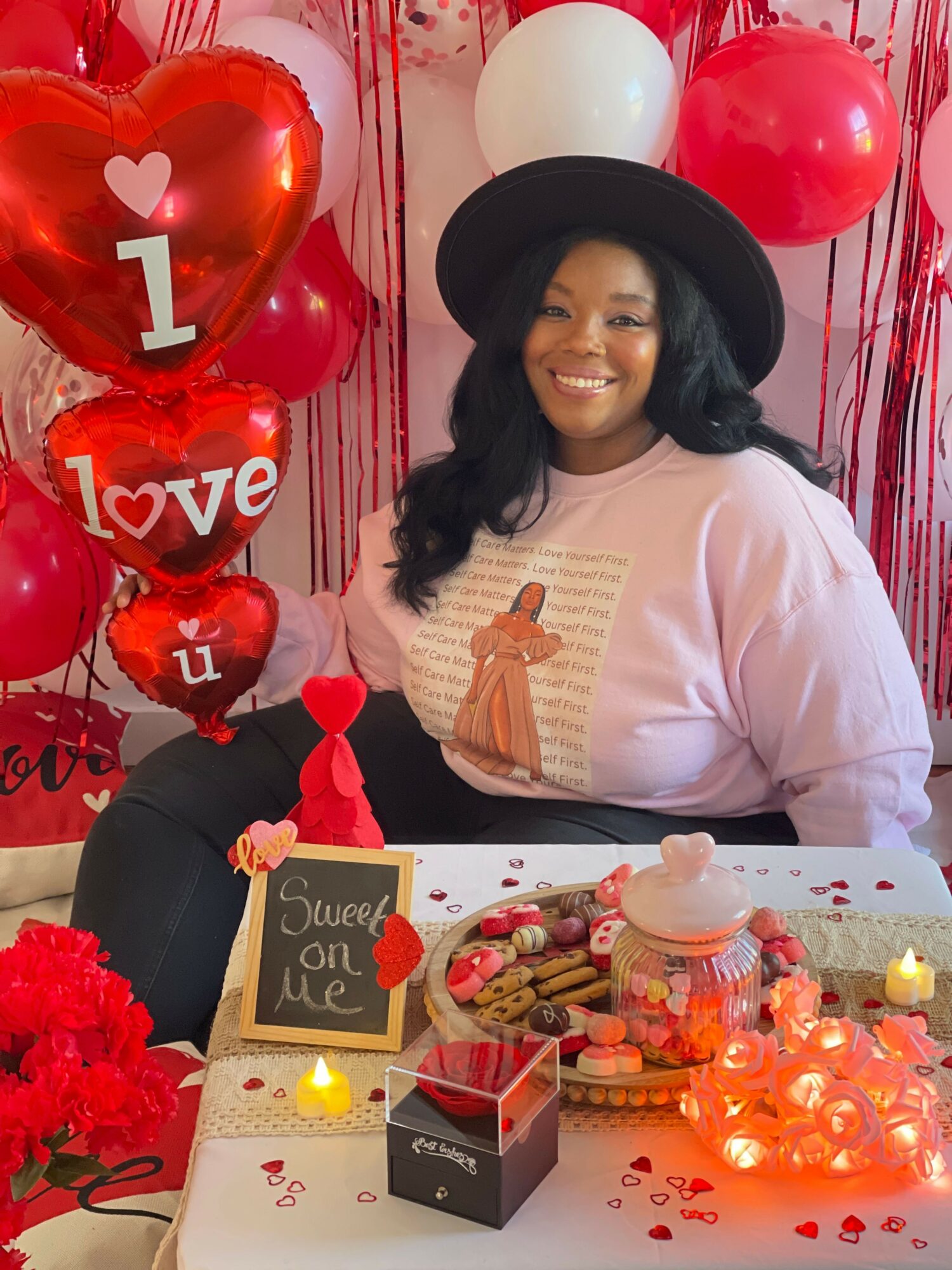 Woman with black hat smiling at a Valentine's Day celebration with balloons, chocolates, and heart decorations.