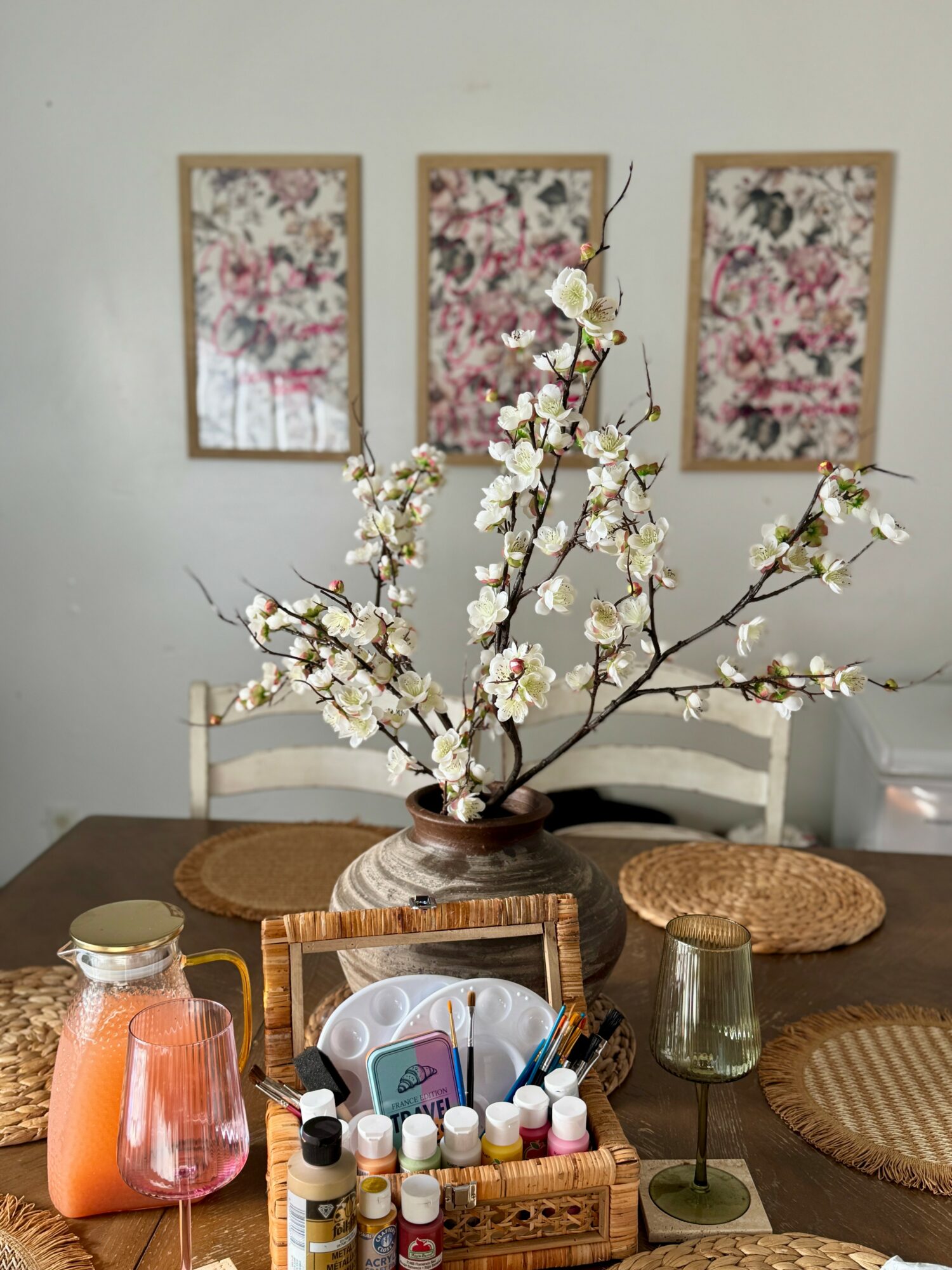 Vase with white flowering branches on a dining table, three woven placemats, and a basket with bottles and a face mask, with framed floral artwork on the wall behind.