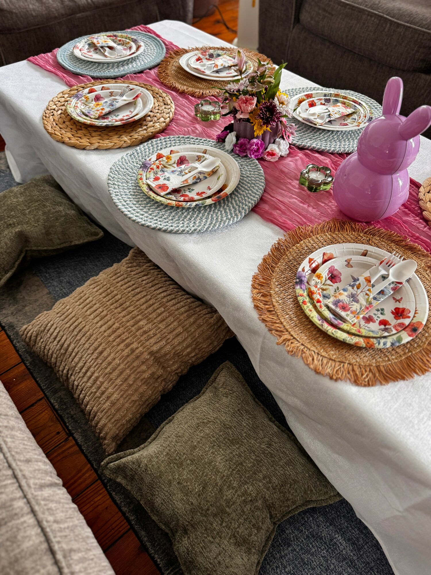 Table with colorful plates, a pink bunny-shaped decoration, and a floral centerpiece, surrounded by cushions and chairs.