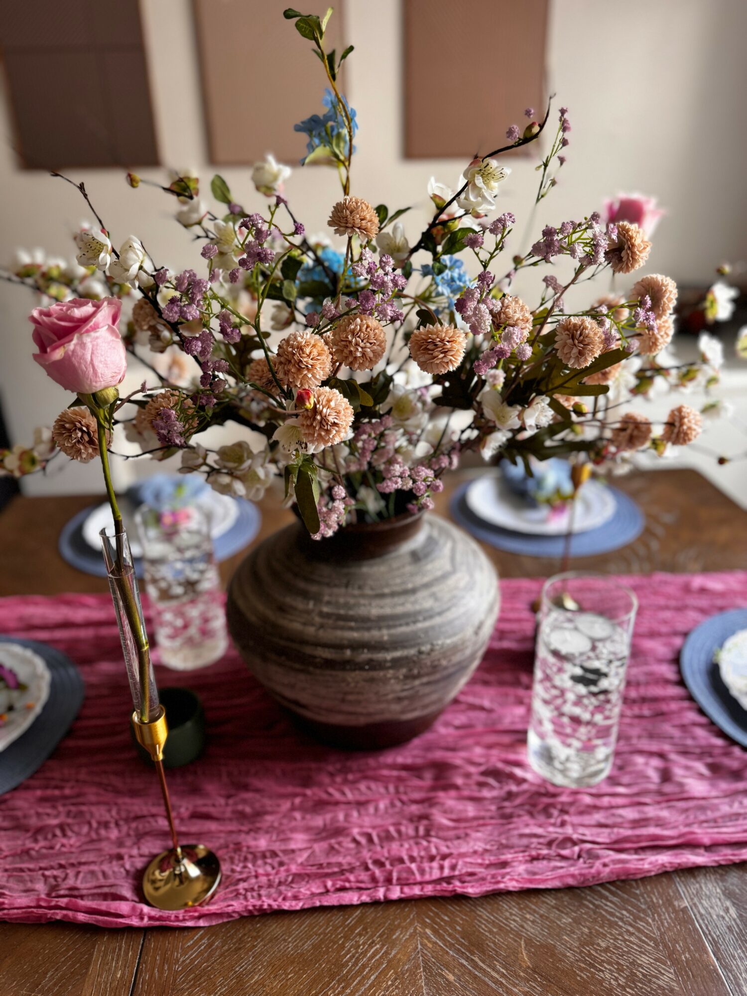 Vase with colorful flowers on a dining table with glasses and plates, pink table runner, and wall art in background.
