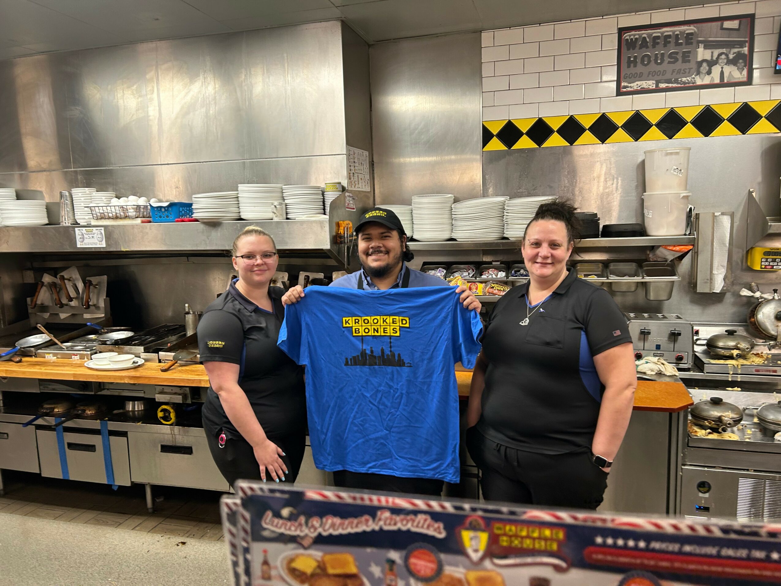 Three people in a kitchen, one holding a blue shirt, with kitchen equipment and shelves in the background.