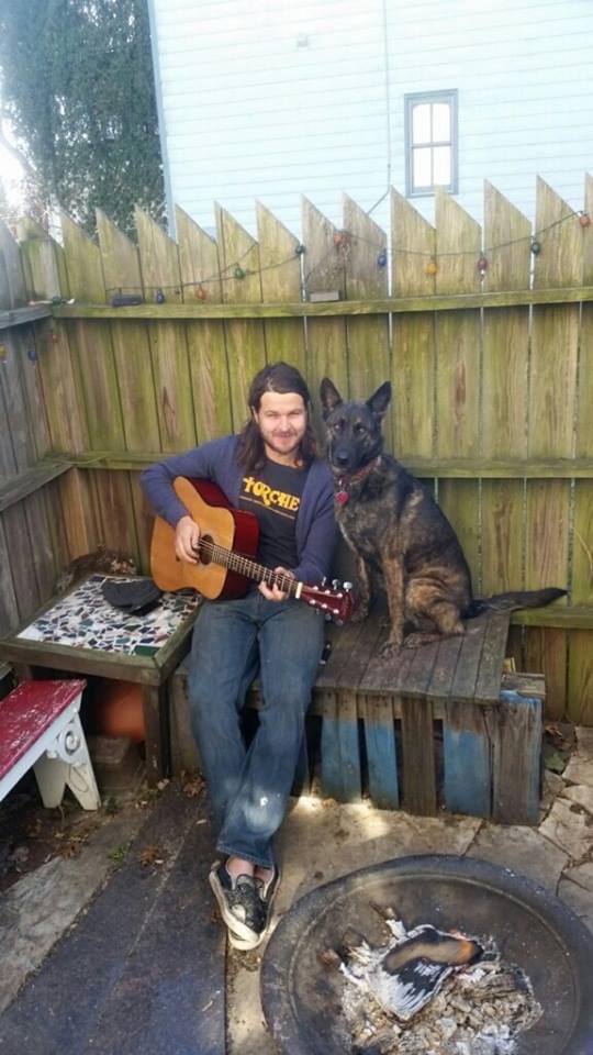 Person sitting on a wooden bench playing guitar with a large dog beside them, in a backyard with a wooden fence.