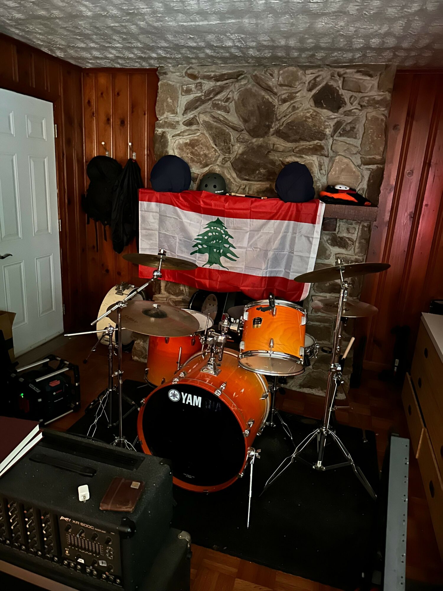 A drum set with orange drums and cymbals on a wooden floor, with a flag featuring a green cedar tree on a white and red background hanging nearby.