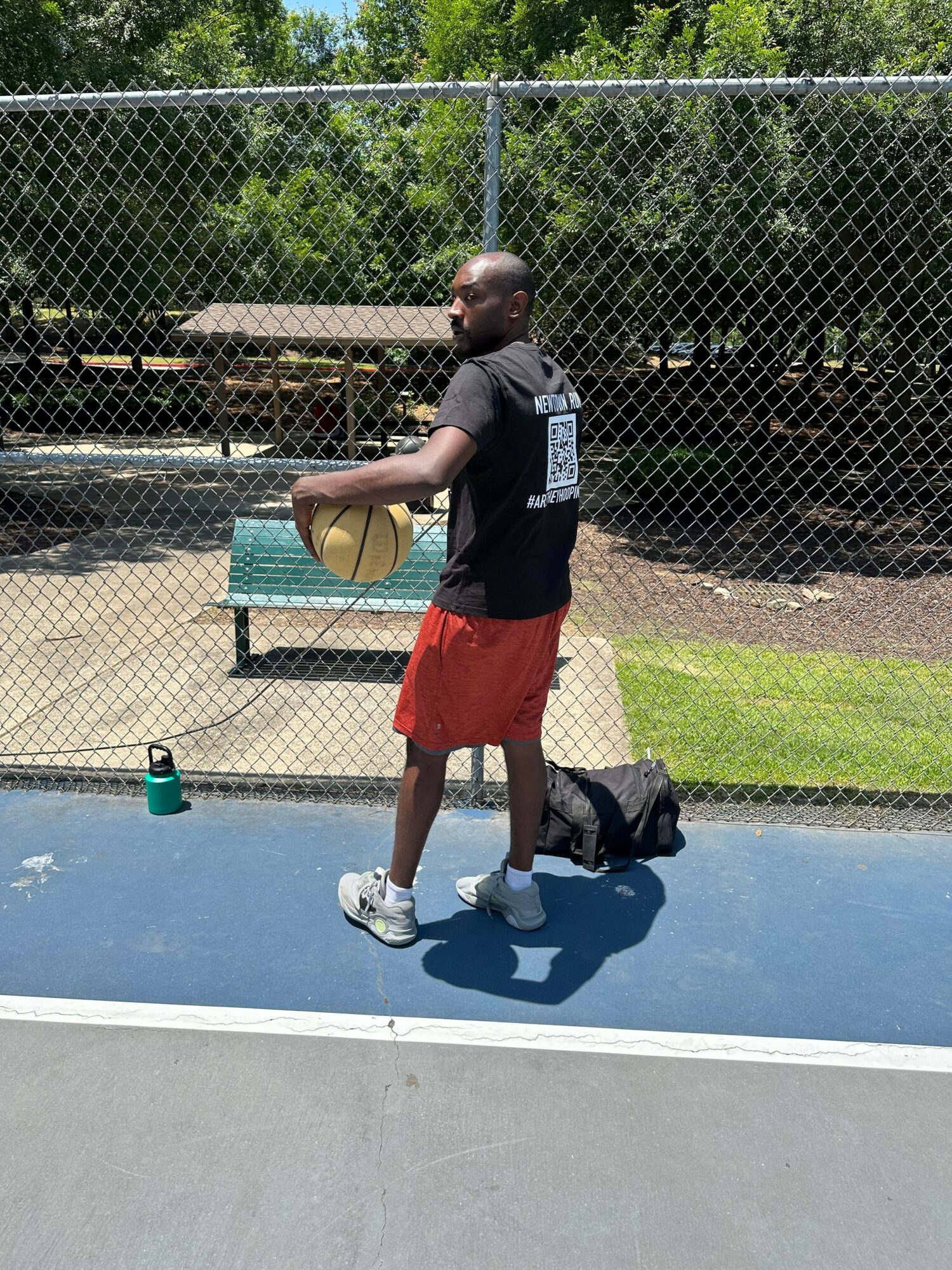 Young man in black shirt and red shorts holding a basketball on a court, with a backpack and water bottle nearby.