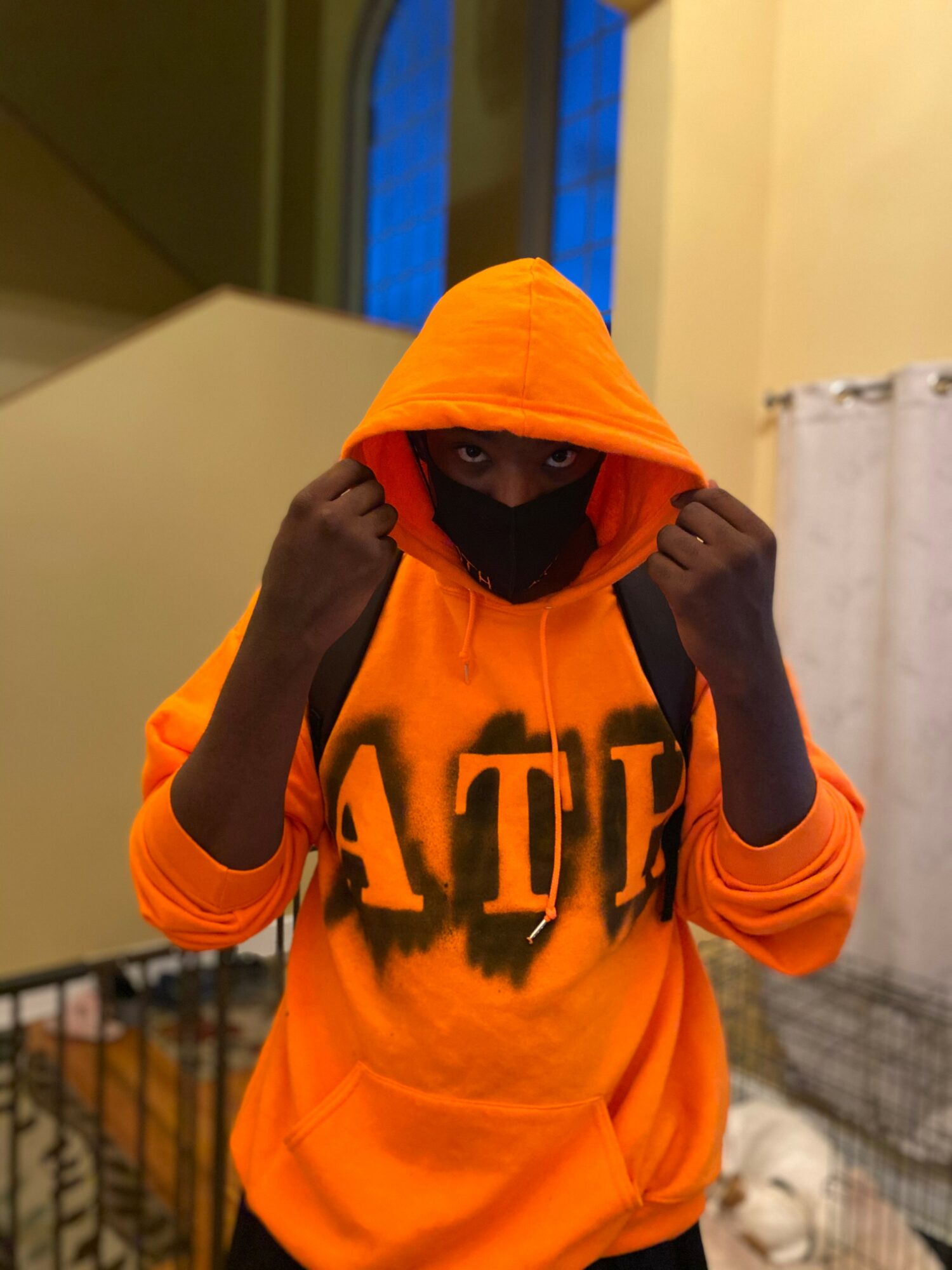 Person wearing an orange hoodie with a black face mask, adjusting the hood, indoors with a window and curtain in the background.