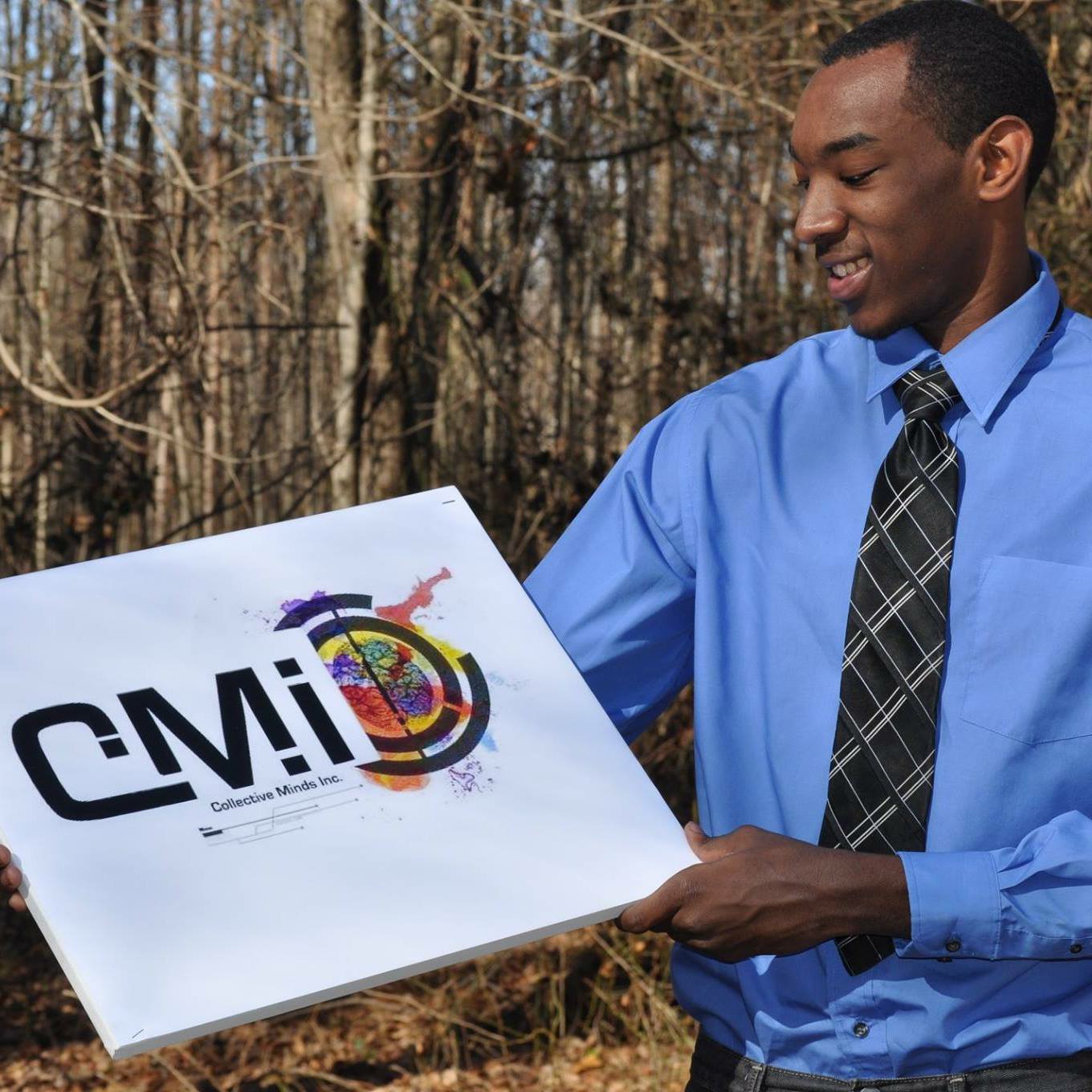 Young man in blue shirt and tie holding a sign with the CMI logo outdoors, trees in background.