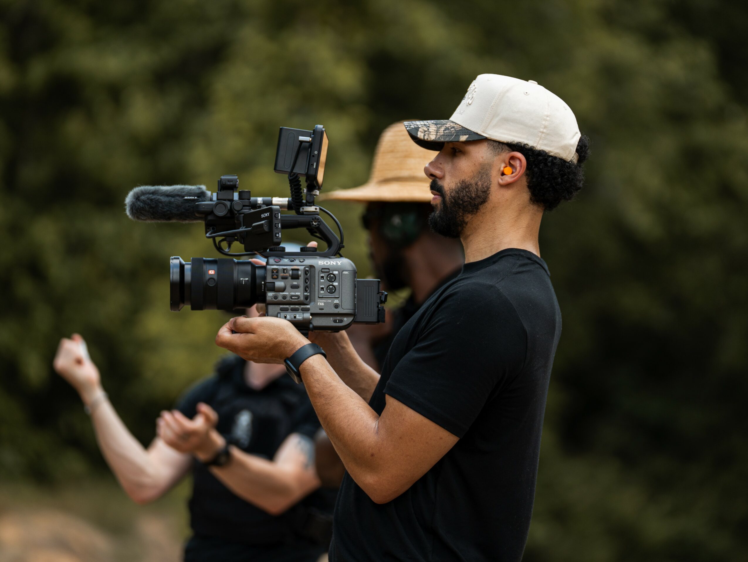 Man with a beard and cap holding a professional video camera outdoors, with a person in the background. Forest in background.