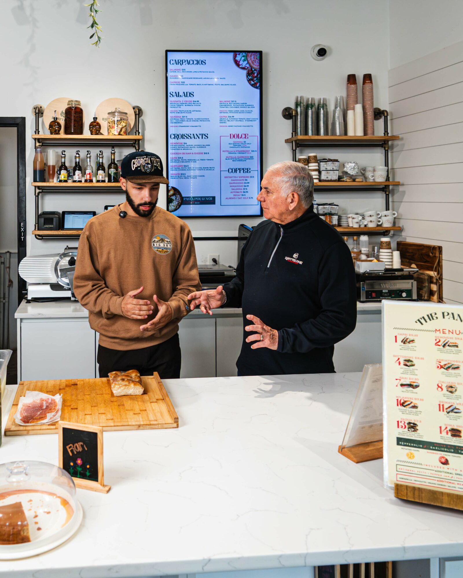 Two men talking inside a cafe, one wearing a brown hoodie and cap, the other in a black jacket, behind a white counter.