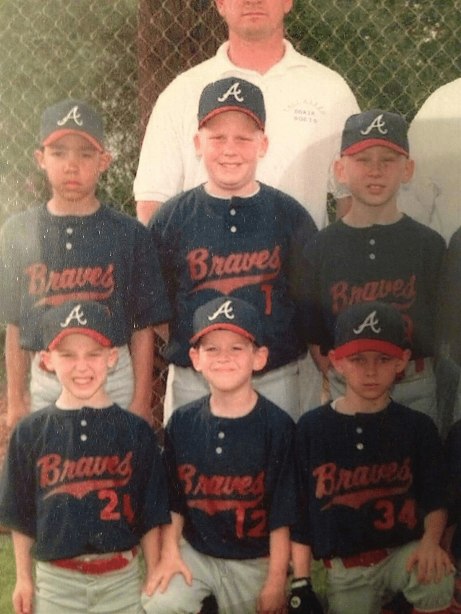 Group of young baseball players and an adult, all wearing Braves uniforms and caps, standing outdoors in front of a fence.
