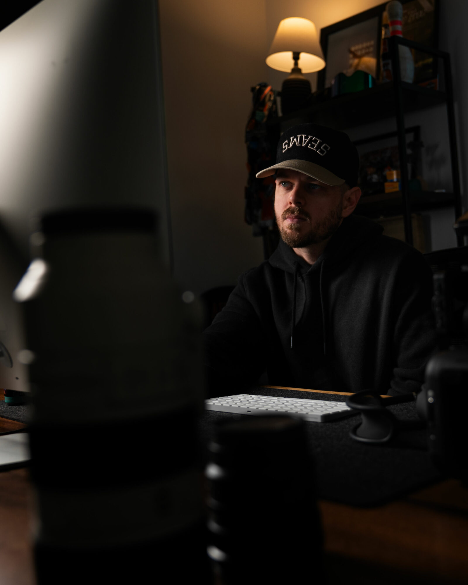 Man wearing a cap and hoodie sitting at a desk in a dimly lit room with a lamp and bookshelf behind him.