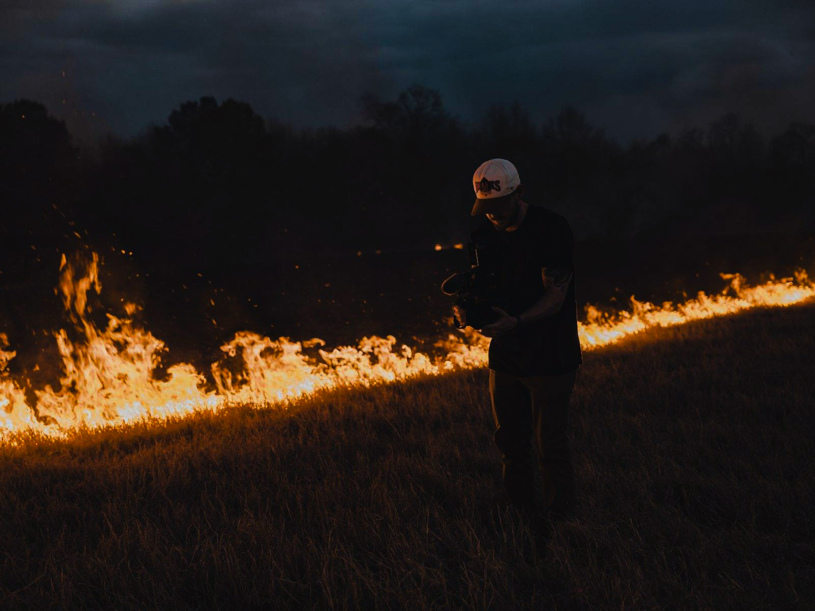Person standing near a line of fire in a field during nighttime, wearing a helmet and dark clothing.