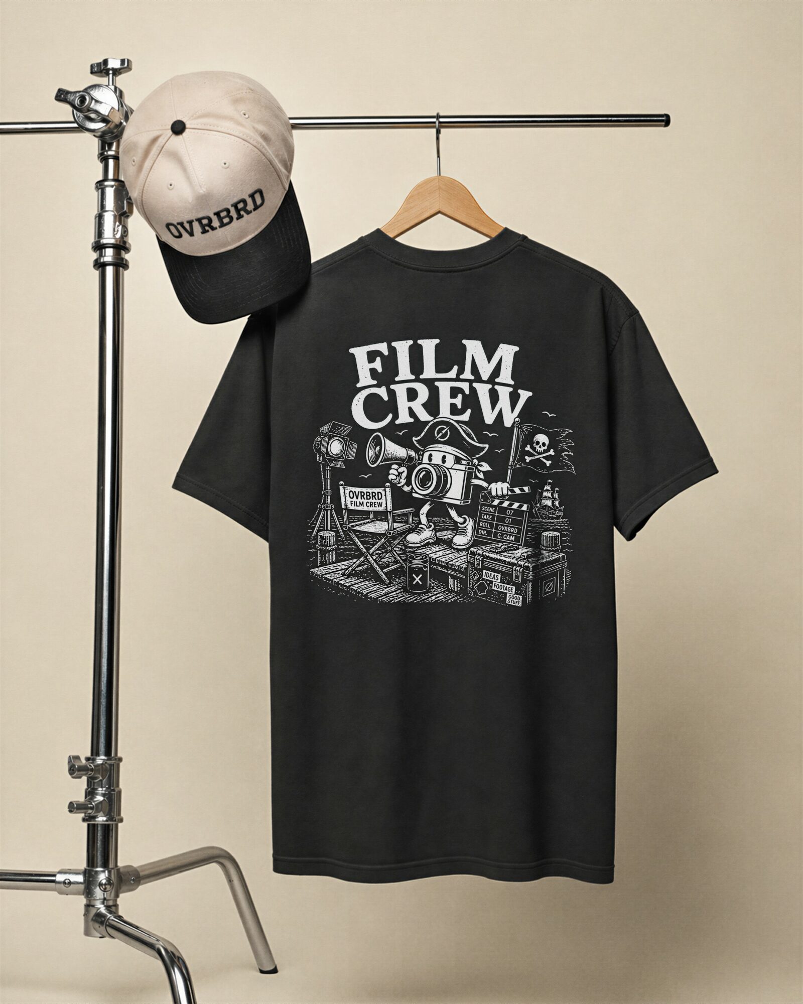 Black T-shirt with film crew graphic hangs on a hanger, with a white and black cap on a metal stand.