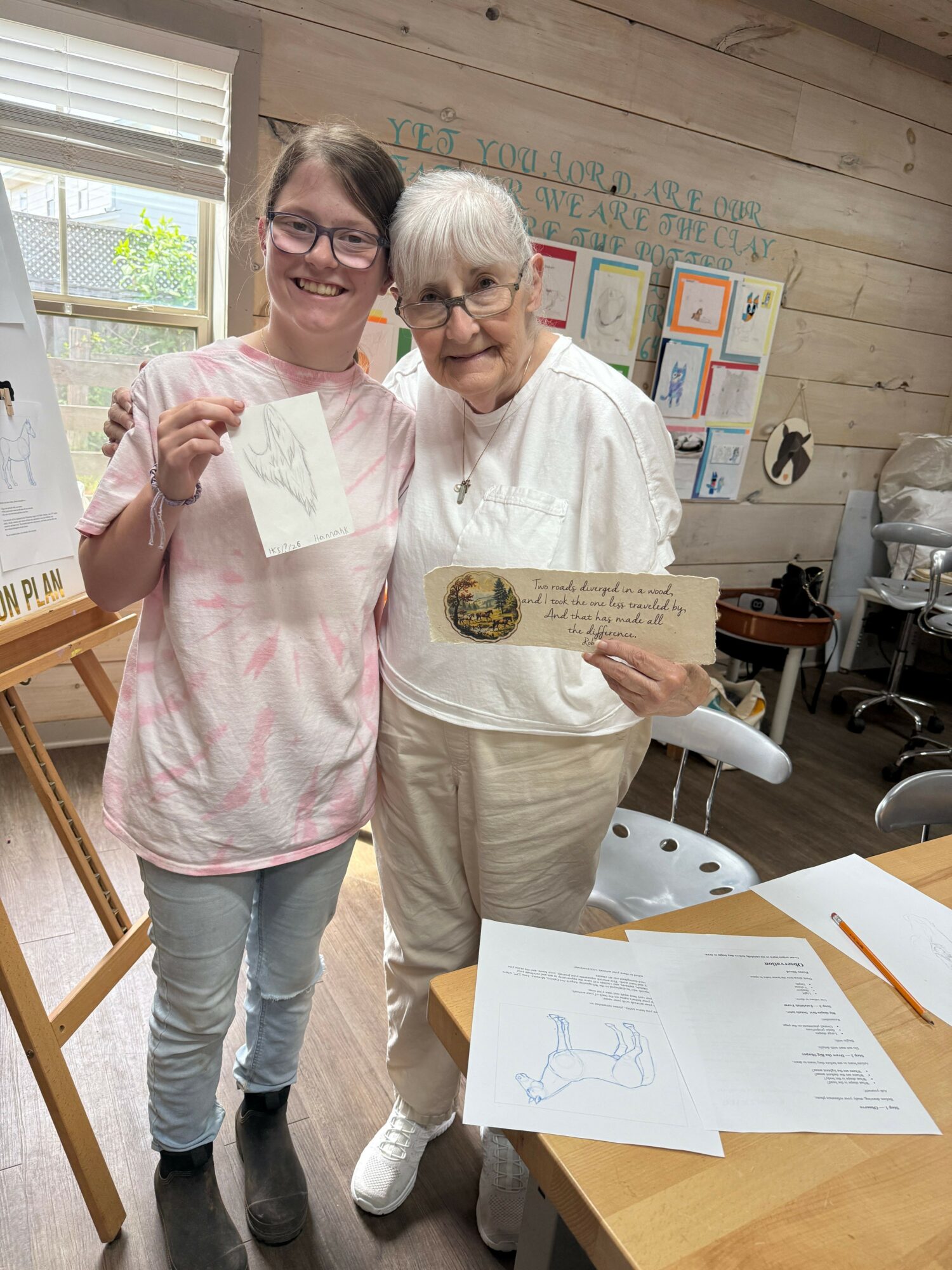 Two women standing indoors, one holding a paper and the other holding a drawing, with a table and window in background.