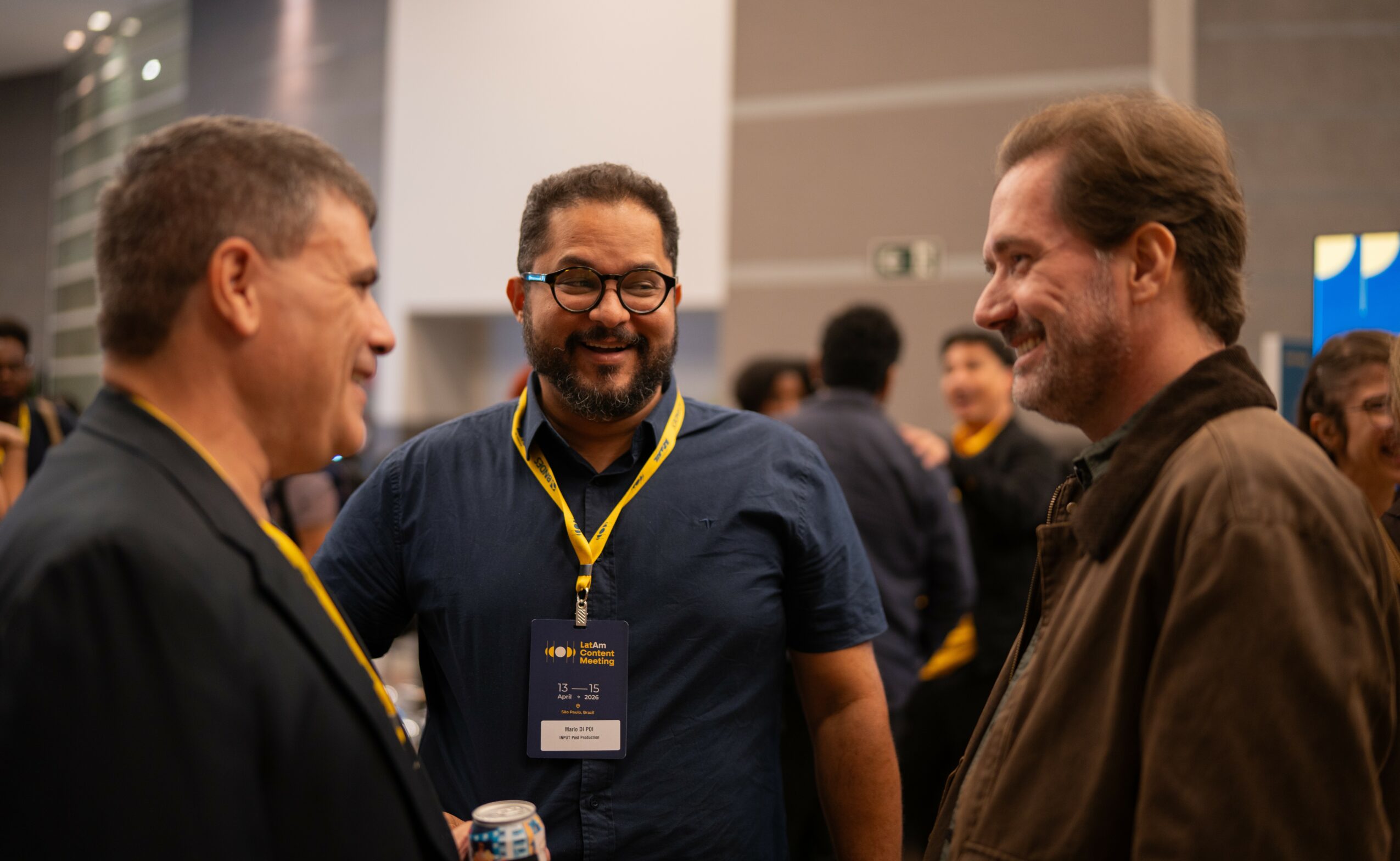 Three men engaged in conversation at a conference, with other attendees in the background.
