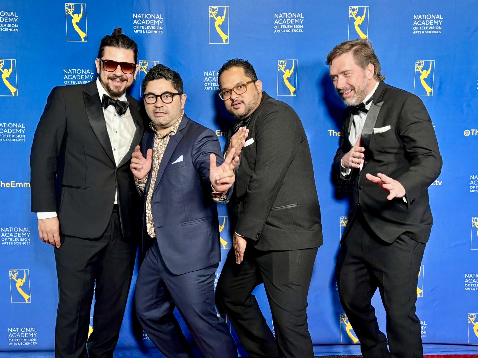 Four men in tuxedos posing on a blue backdrop with logos, smiling and making gestures at an event.