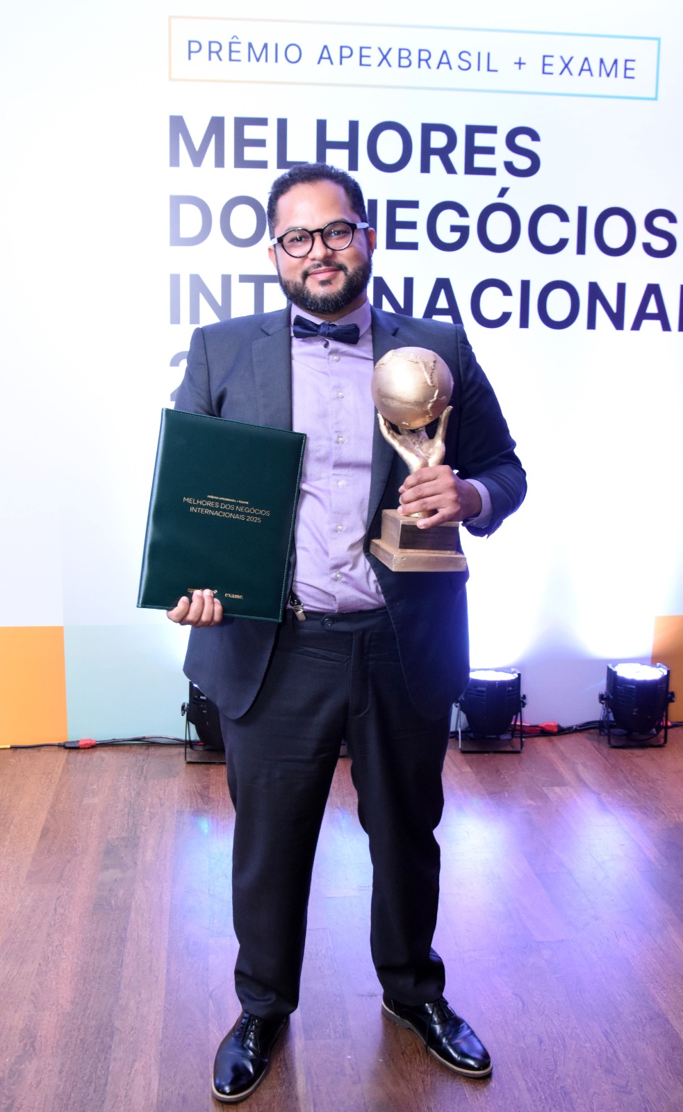 Man in suit holding a trophy and a folder, standing on stage with a white background and text, smiling.