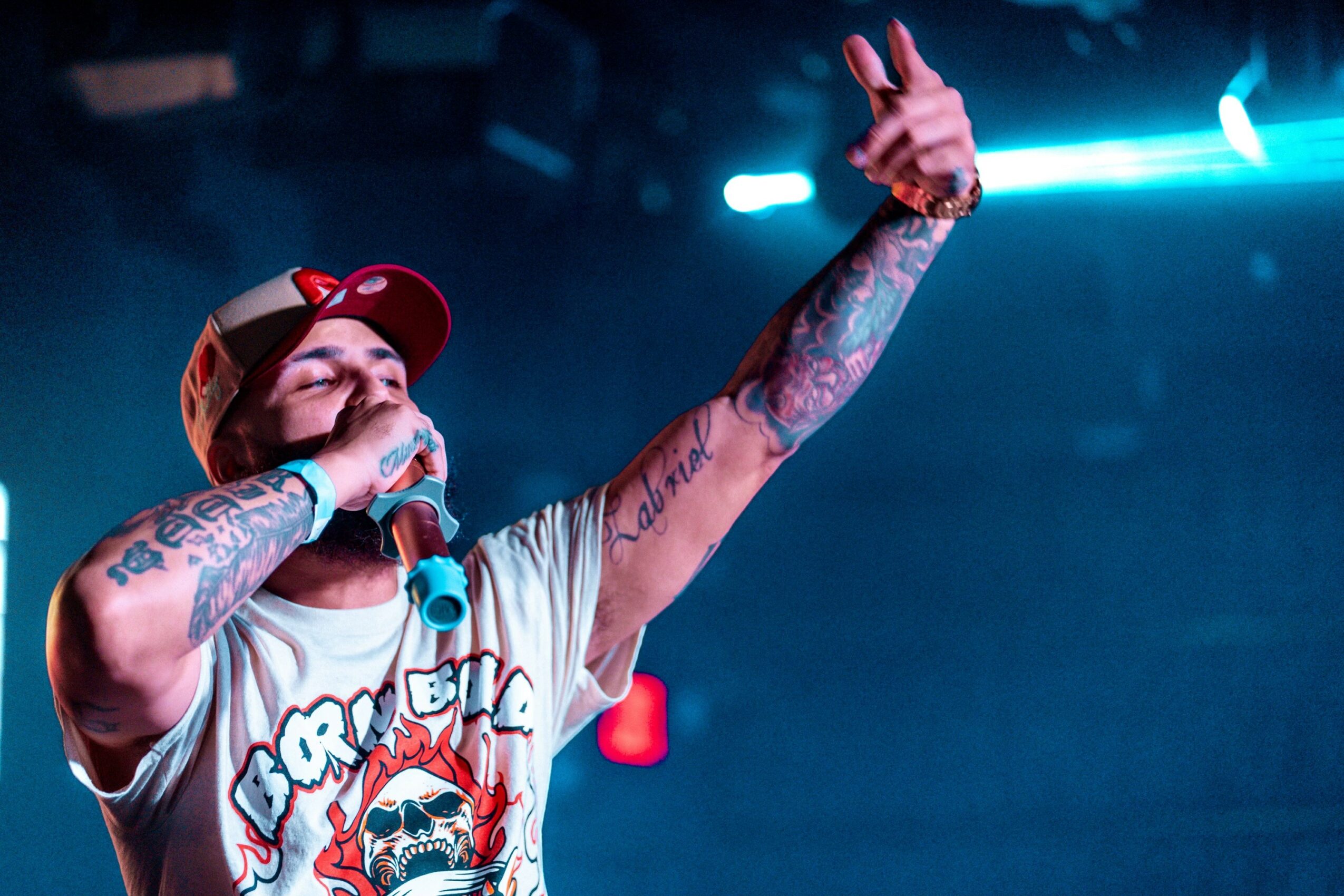 Person singing into microphone with tattoos, wearing a cap, on stage with blue lighting