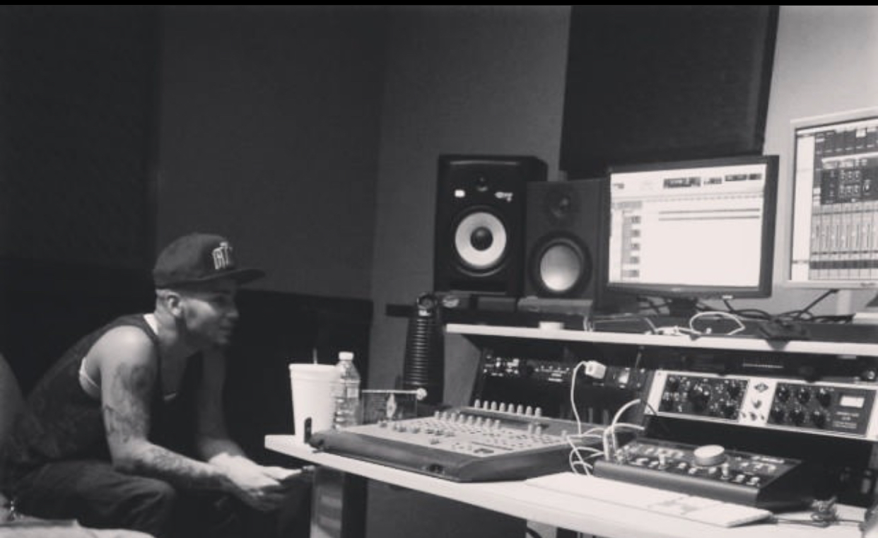 Person with tattoos wearing a cap in a recording studio with audio equipment and monitors.