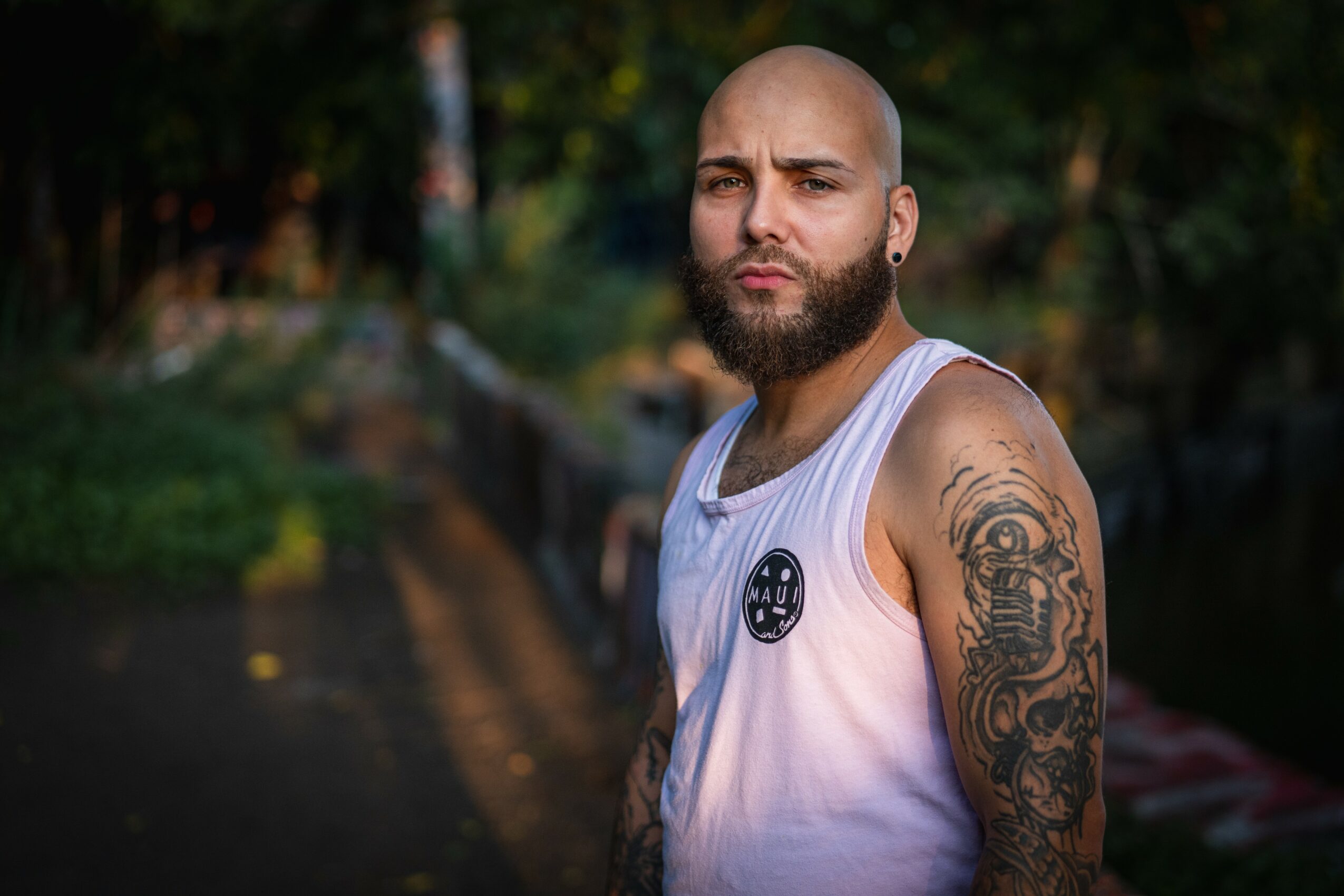 Bald man with beard and tattoos on arm, wearing a white sleeveless shirt, outdoors with trees and pathway in background.
