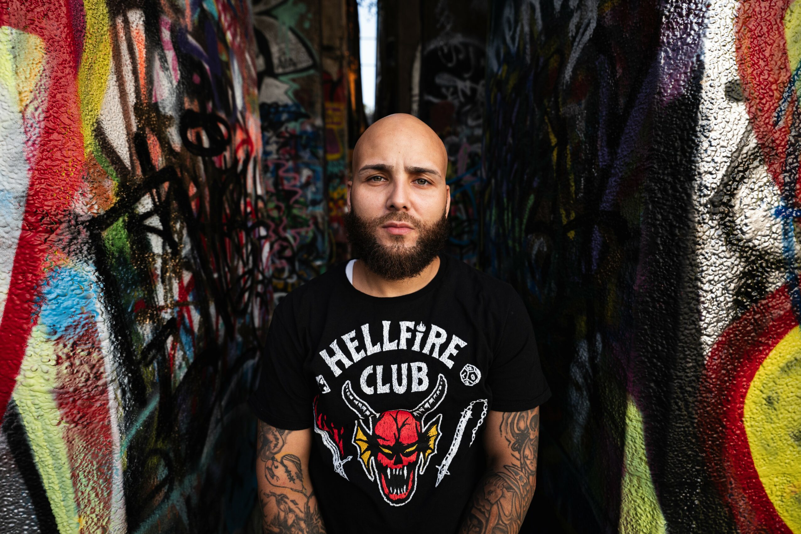 Bald man with a beard and tattoos standing in front of a colorful graffiti wall.