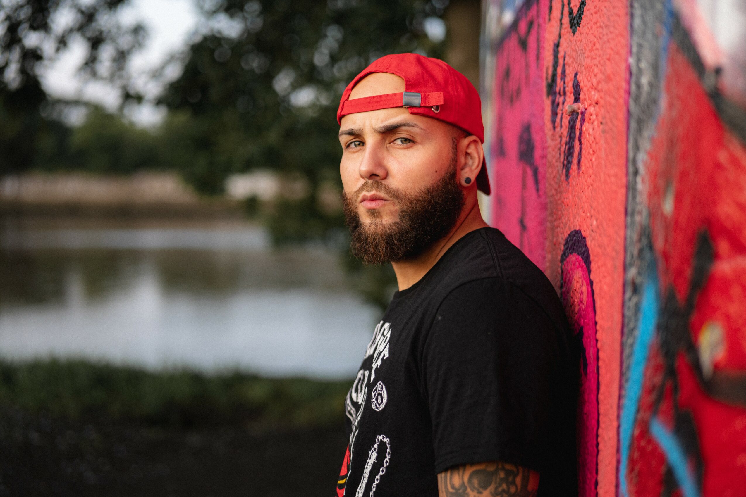 Young man with a beard wearing a red cap backwards, leaning against a colorful graffiti wall near a body of water.