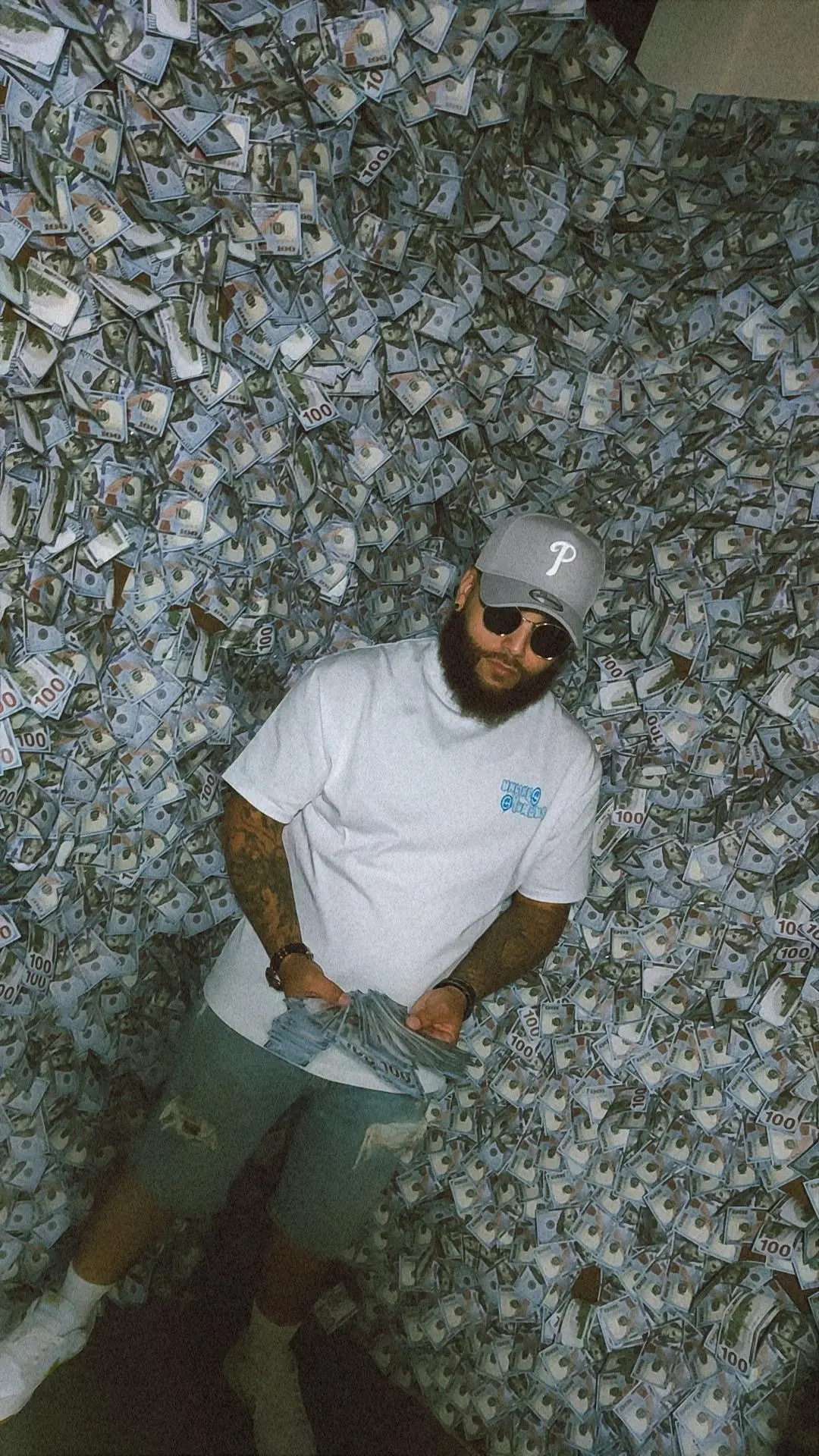Person with sunglasses and a cap stands in front of a wall covered with numerous dollar bills.