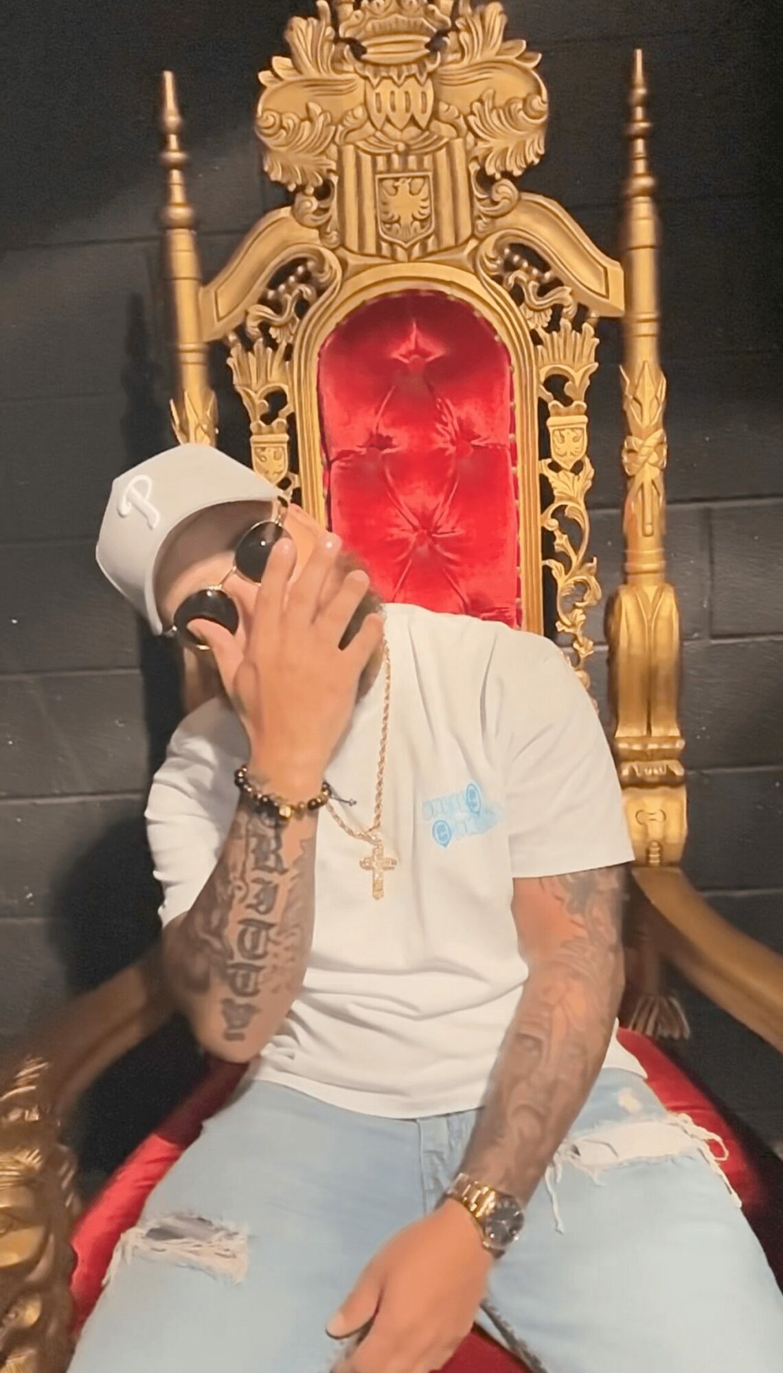 Person sitting on a throne with a red cushion, wearing a white cap and shirt, holding a phone to their face.