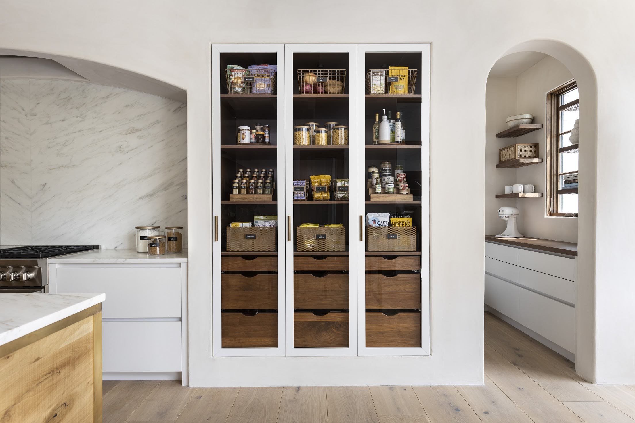 Three tall pantry cabinets with open shelves and wooden drawers, in a kitchen with white walls and light wood flooring.