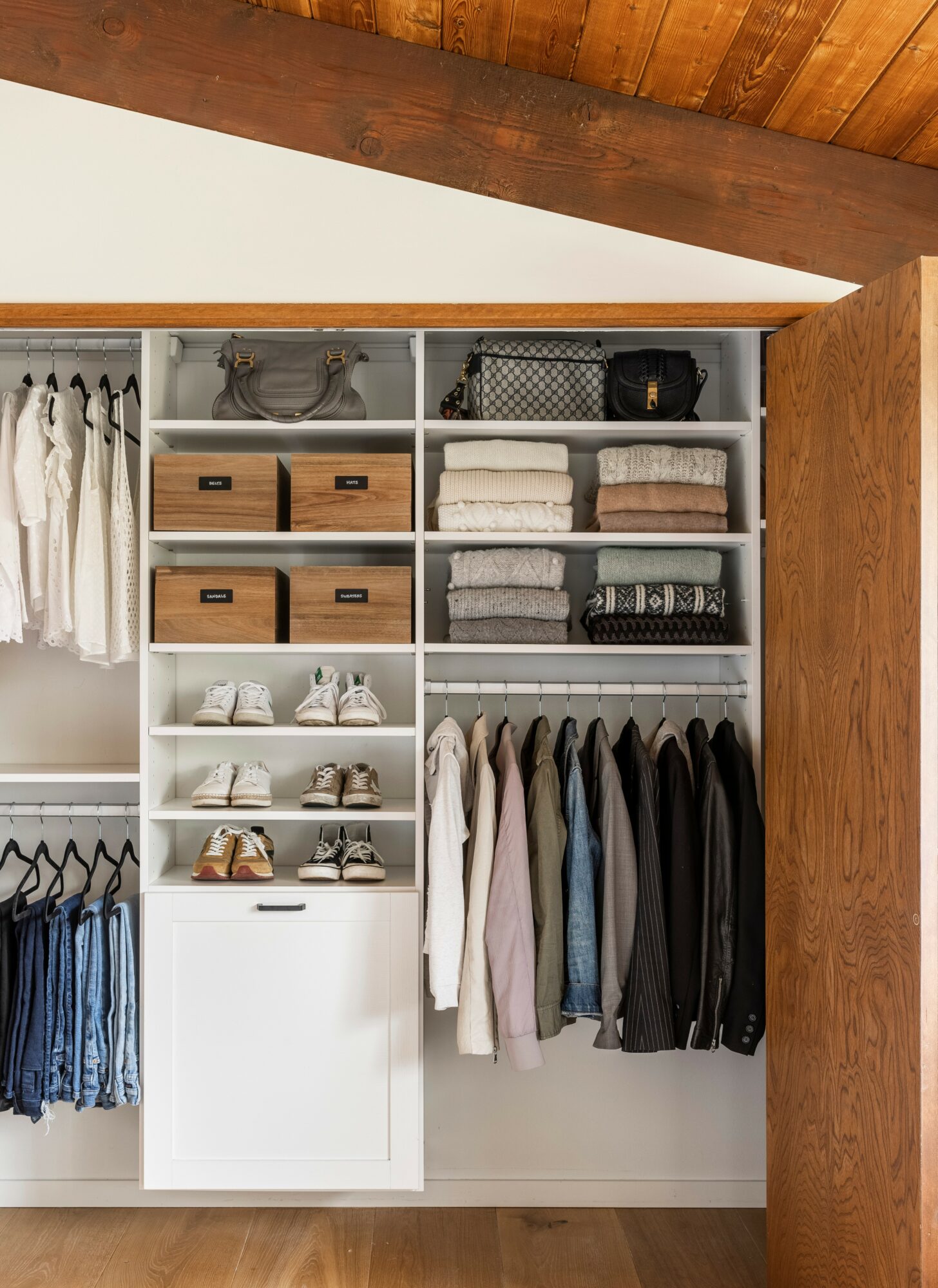 Open closet with shelves, drawers, and hanging clothes, under a wooden ceiling. Clothes are folded and hung neatly.