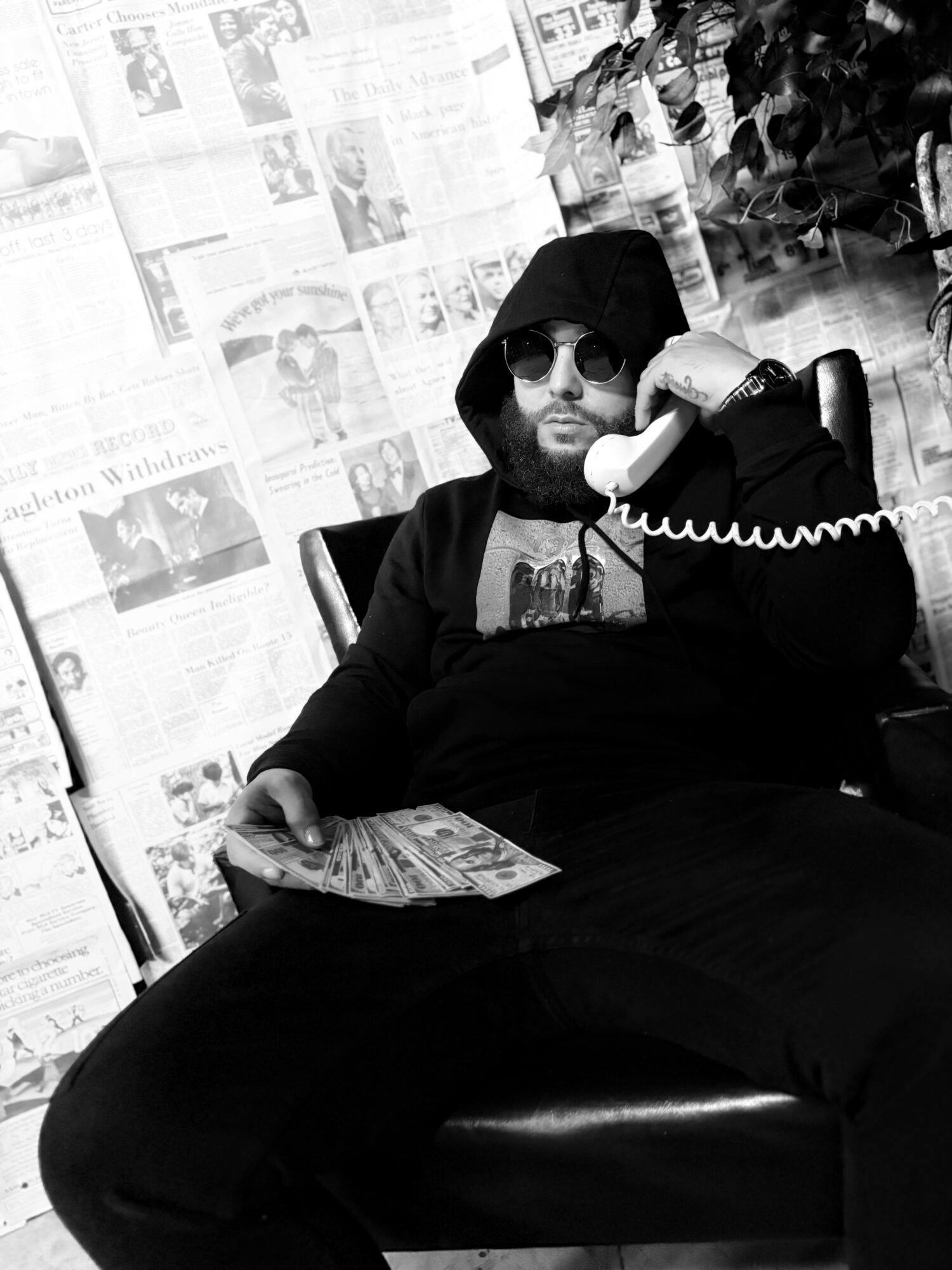 Man with sunglasses and hoodie sitting on a chair, holding a telephone receiver, with newspapers in the background.