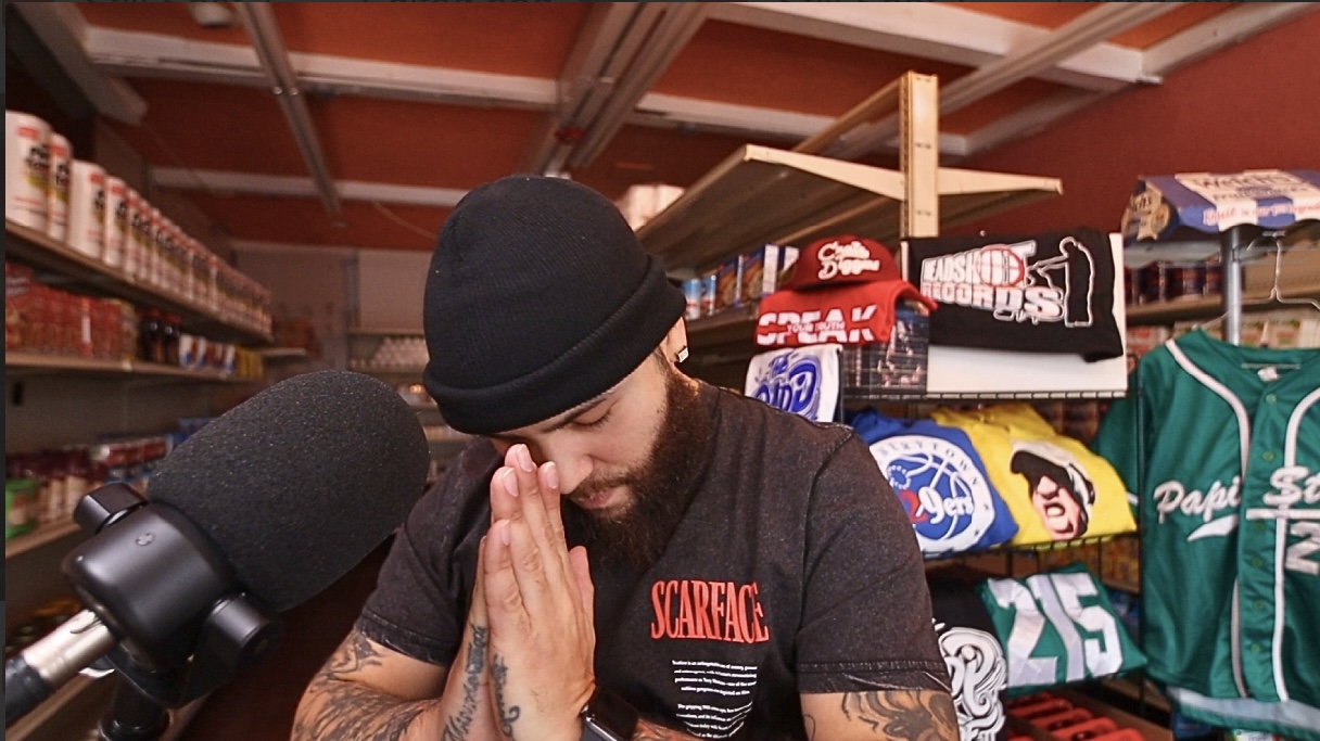 Person wearing a black beanie and black T-shirt with hands pressed together in front of a microphone, surrounded by shelves of clothing and accessories.